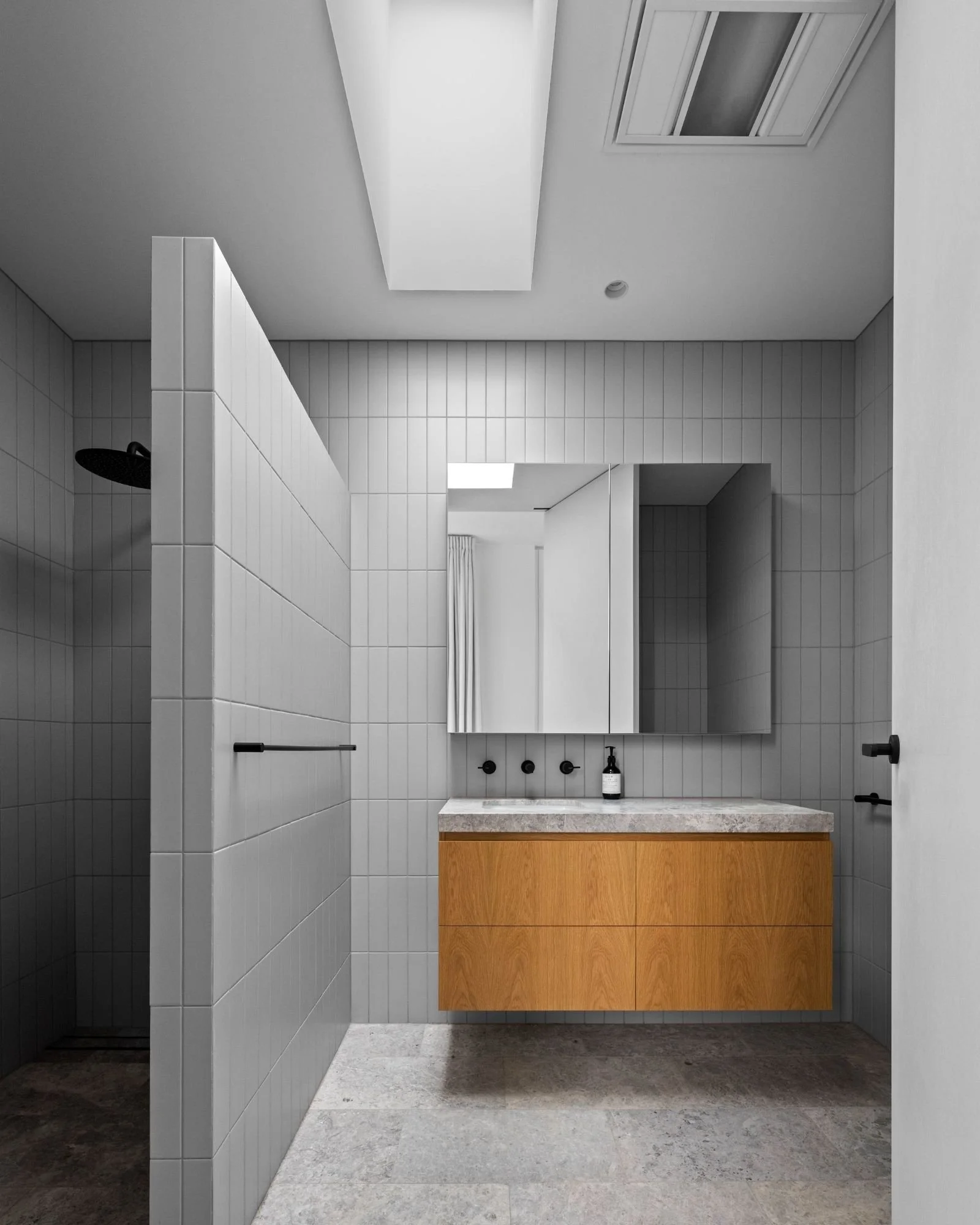 Modern bathroom with large mirror, white tiled walls, black fixtures, a wooden vanity with a beige countertop, and a bottle of soap or lotion on the counter.