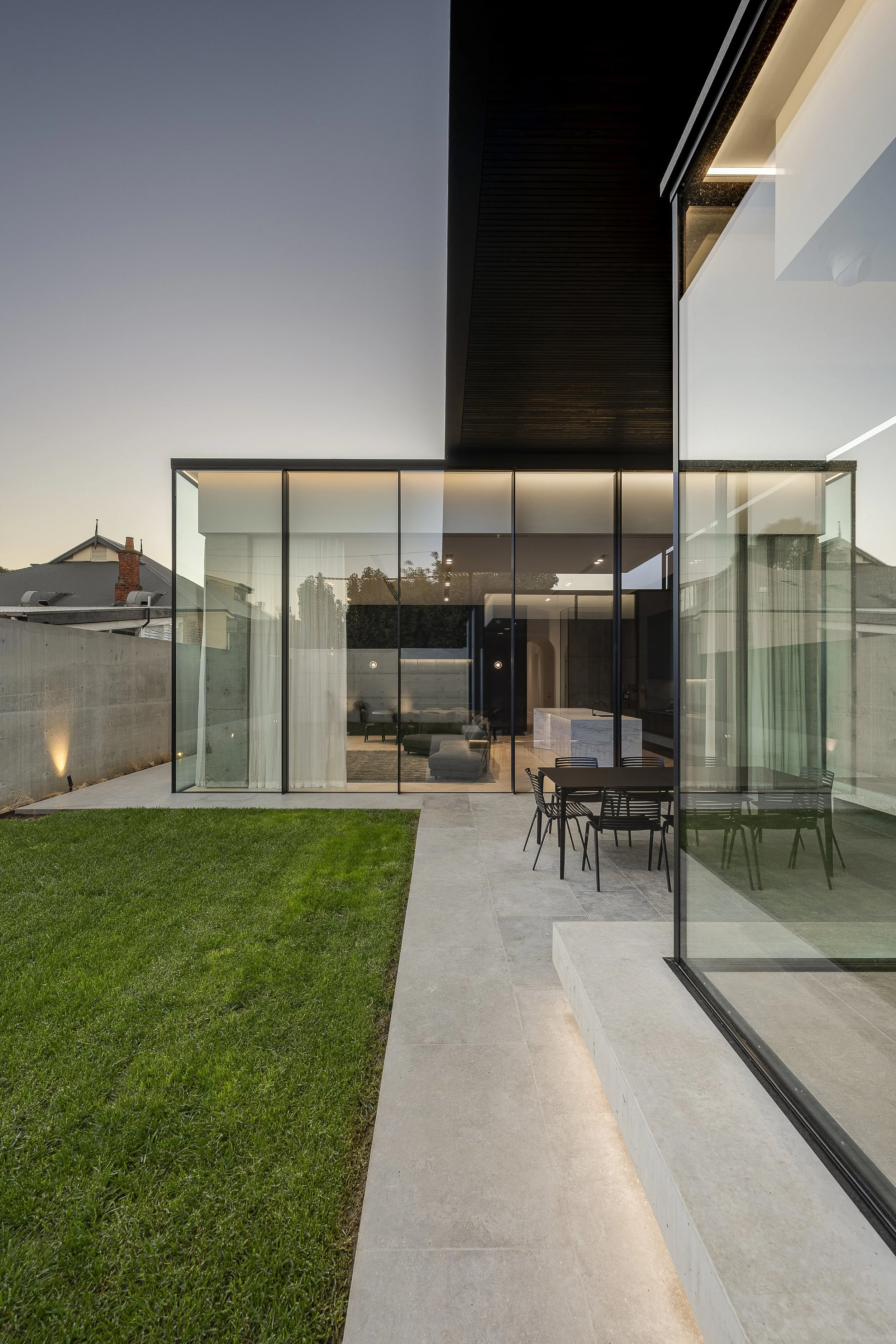 Modern house with glass walls, backyard patio with dining table and chairs, green grass lawn, and neighboring houses in the distance at dusk.