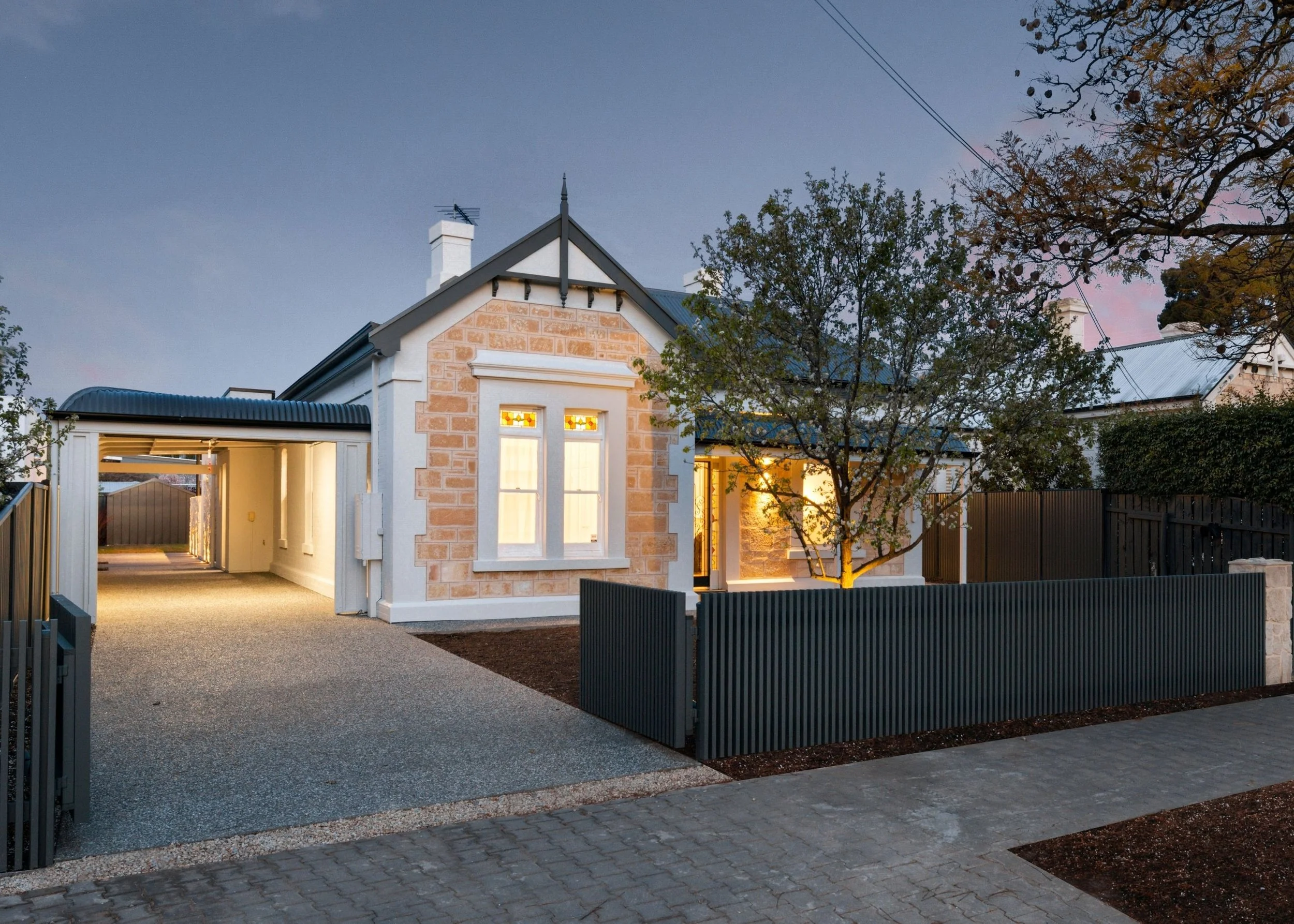 A modern house with a gated driveway, illuminated windows, and surrounding trees during twilight.