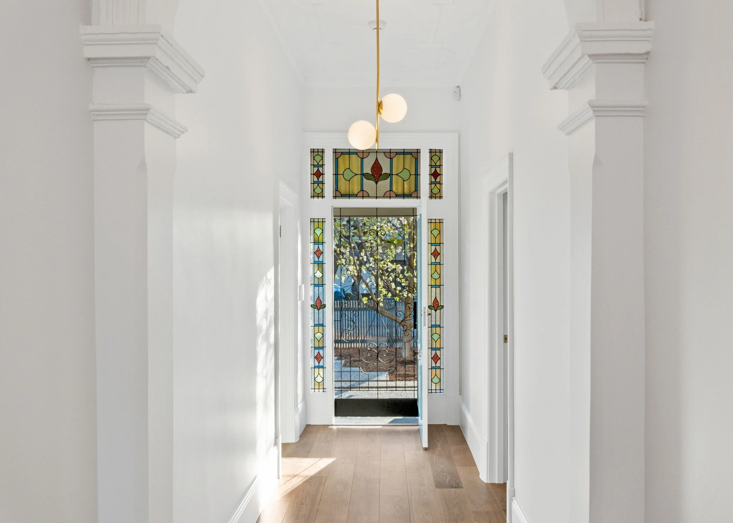 A hallway with a stained glass door and window, white walls, and wooden flooring, with sunlight casting shadows.