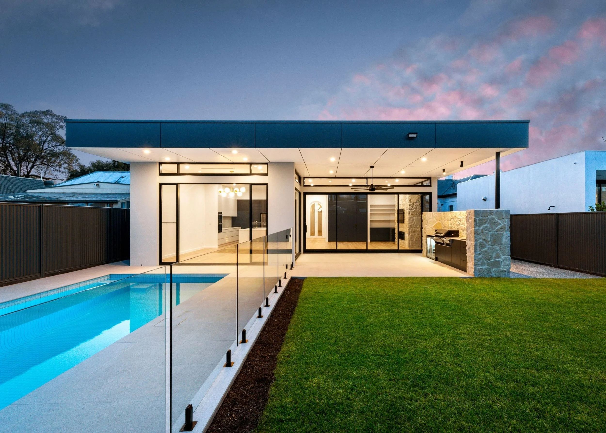 Modern house with a glass sliding door, a swimming pool, a lawn, and an outdoor barbecue area, under a blue sky at dusk.
