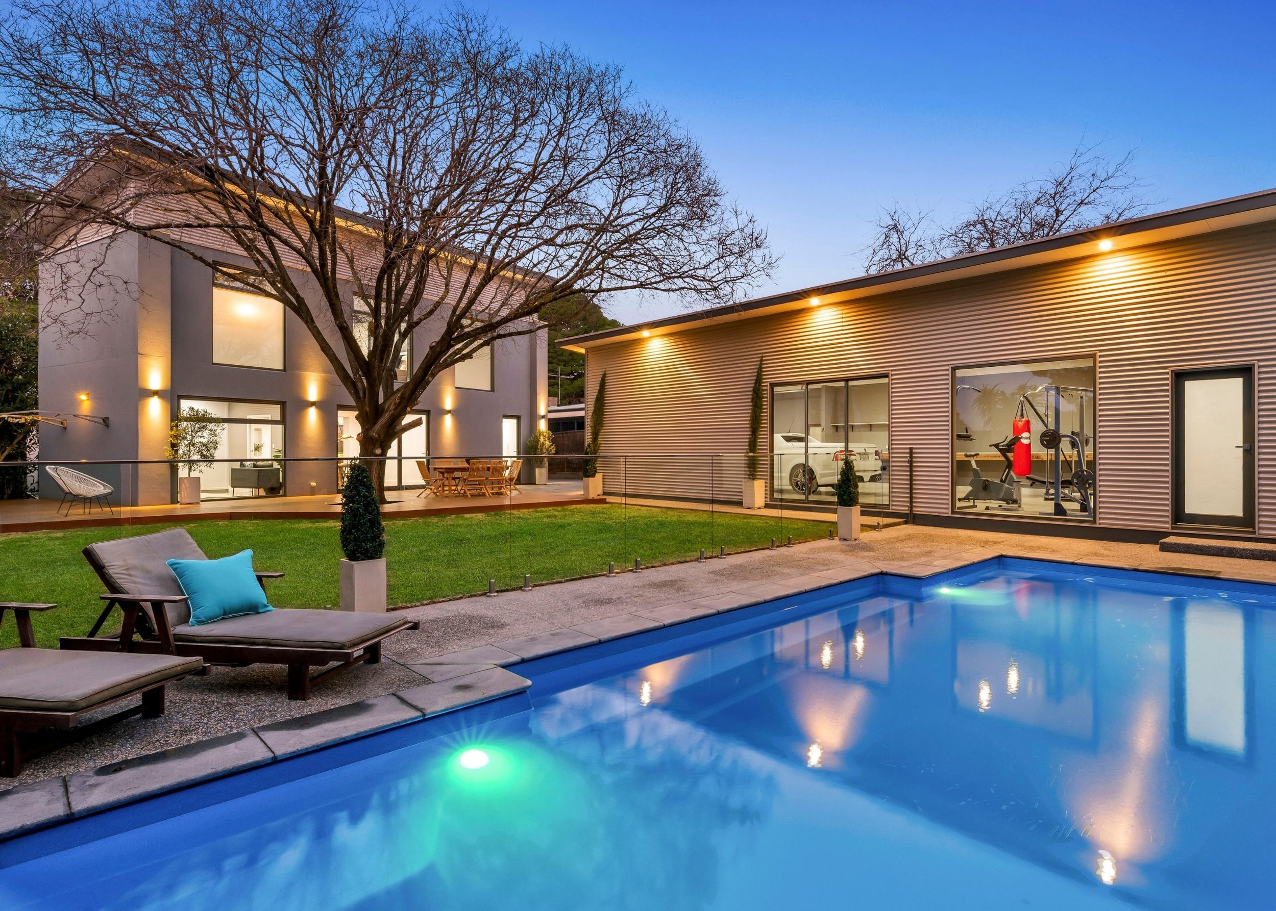 Modern house with large windows, and an outdoor patio with chairs and a table, a backyard swimming pool with illuminated lights, a leafless tree, and a fitness room with gym equipment visible through glass windows, during dusk.