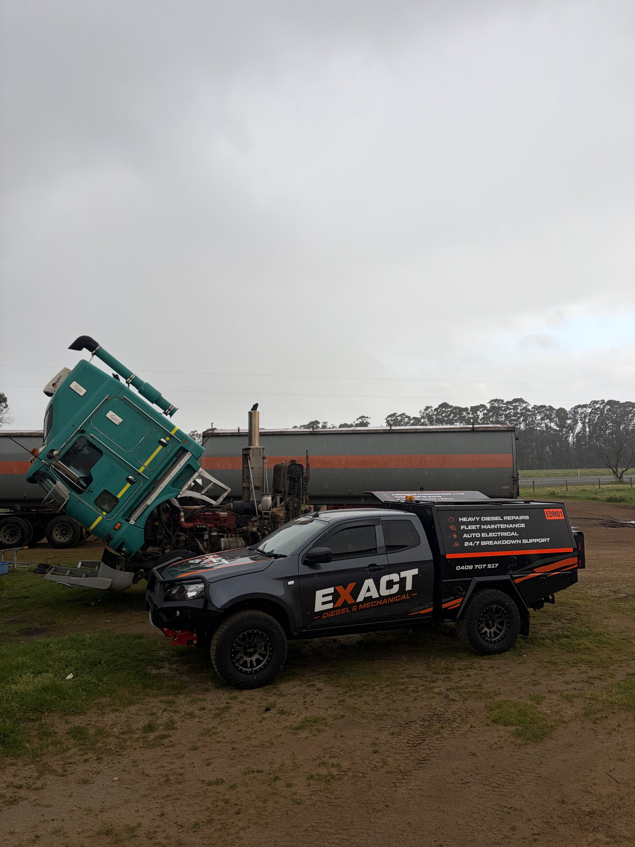 Black Exact Diesel & Mechanical service truck parked beside a damaged green truck and heavy-duty trailer at an industrial repair yard.