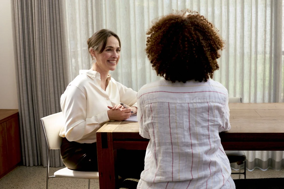 Two women having a conversation at a table, one facing the camera and smiling, the other with back to the camera, in a room with curtains