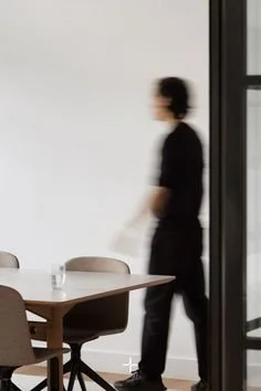 A person standing near a window in a minimalist room with a table and chairs.
