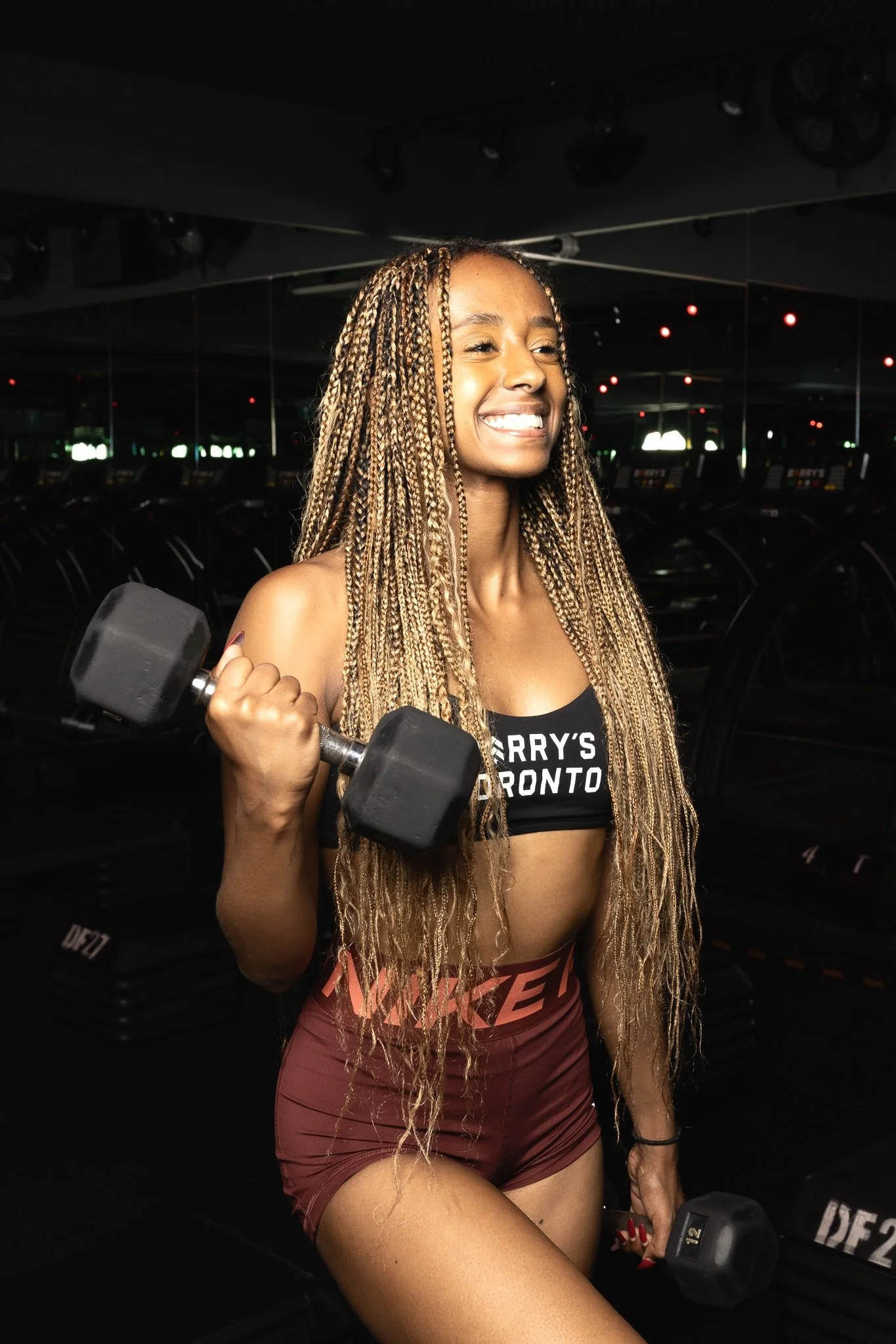 Smiling woman with long blonde box braids lifting dumbbells in a dark gym