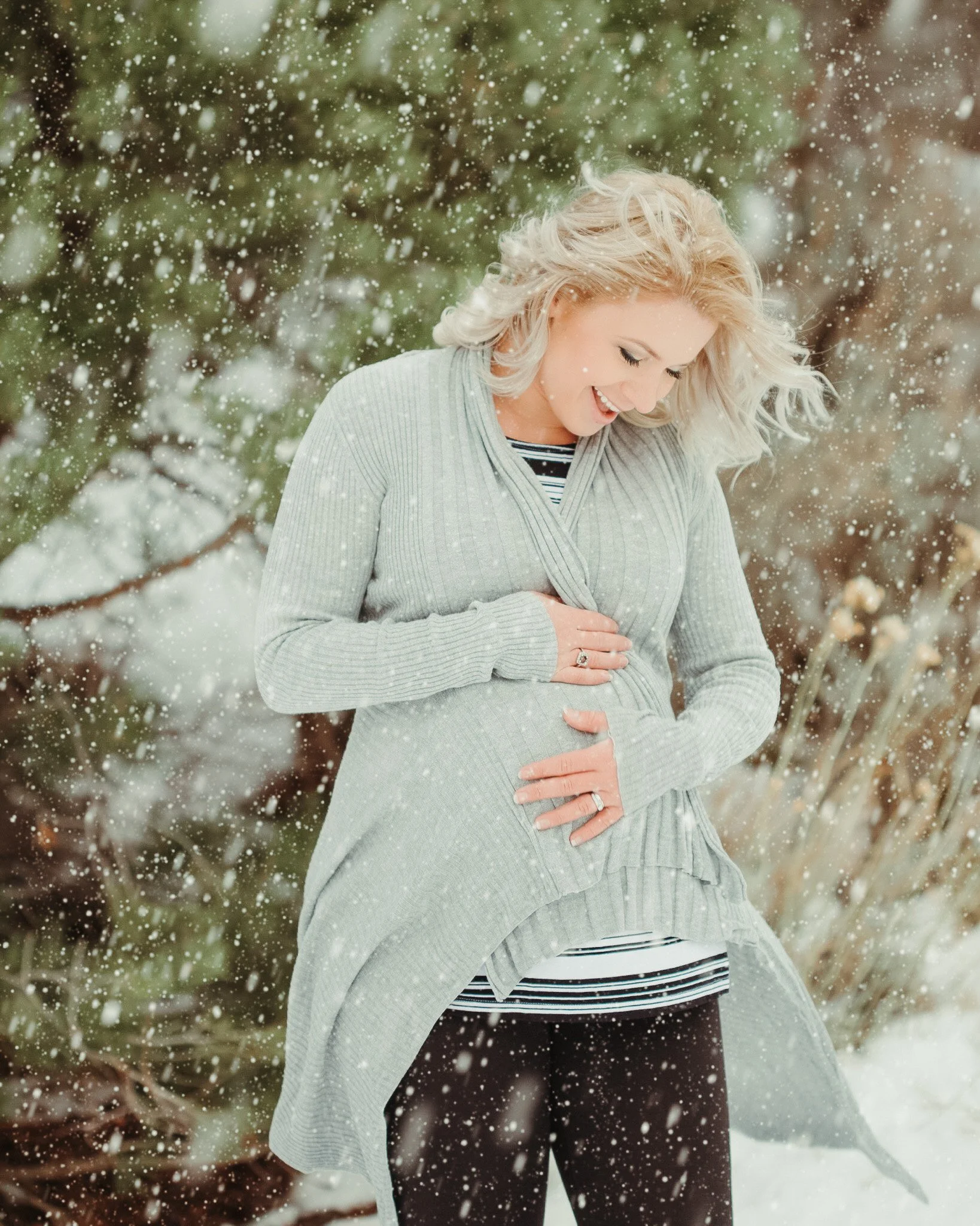 A woman smiling outdoors in a snowy setting, holding her pregnant belly, wearing a gray cardigan over a striped shirt.