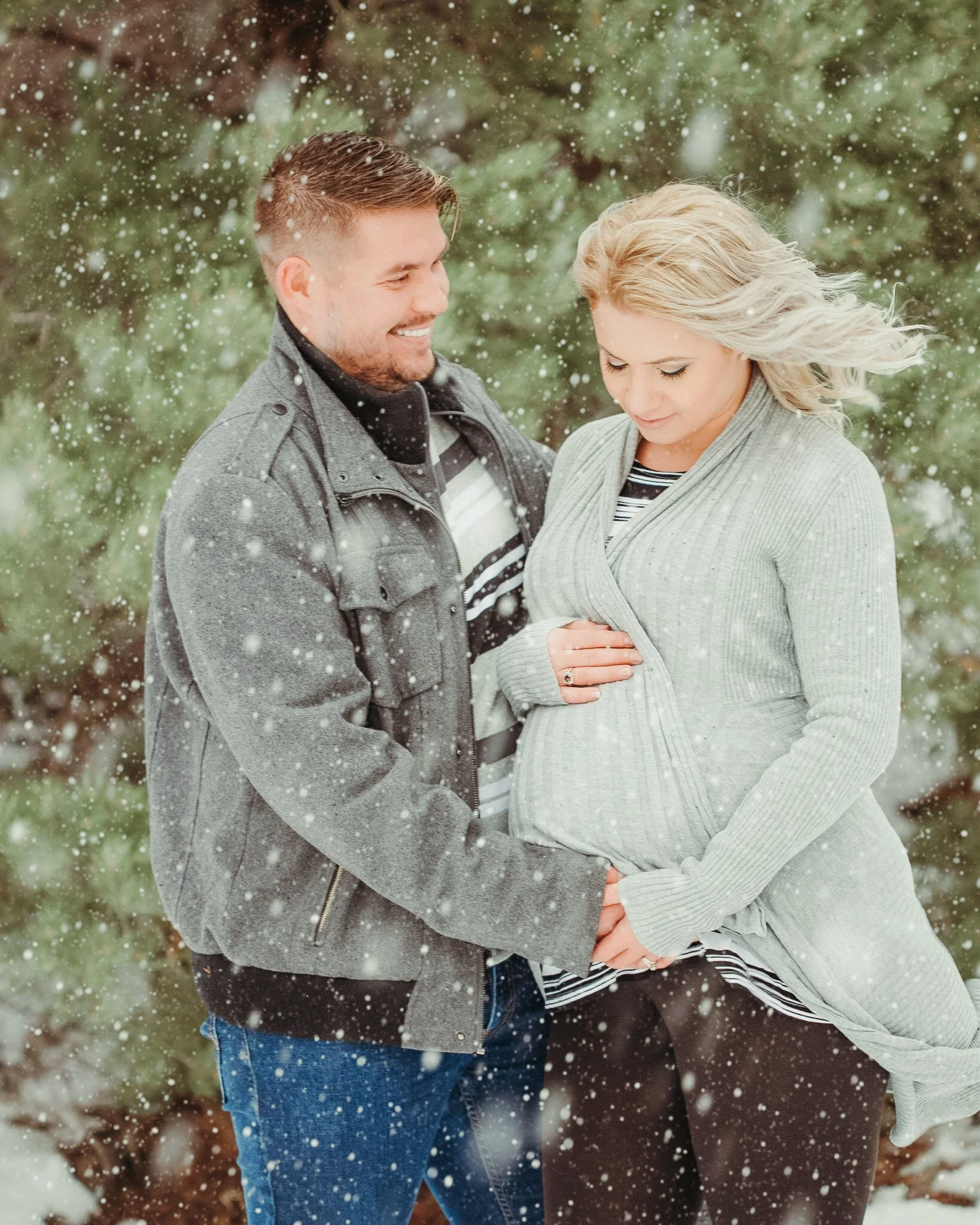 A close-up of a couple holding hands outdoors in a snowy environment with evergreen trees in the background, during winter.