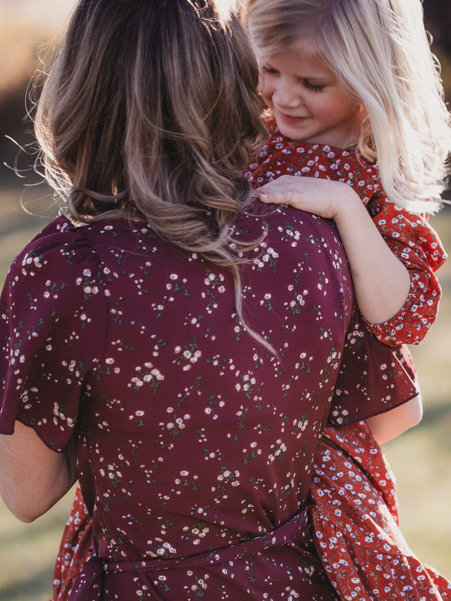 mother in burgundy floral dress holding daughter in red floral dress smiling at each other