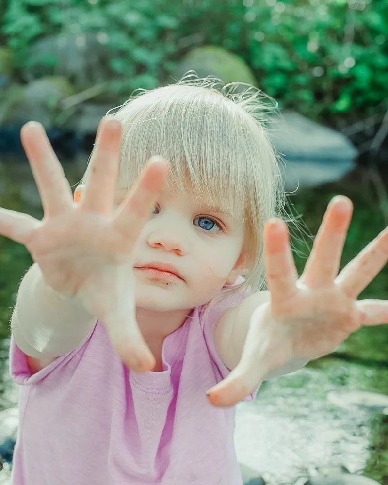 A young girl with blonde hair and blue eyes reaching out towards the camera with both hands, outdoors near a stream with rocks and green foliage
