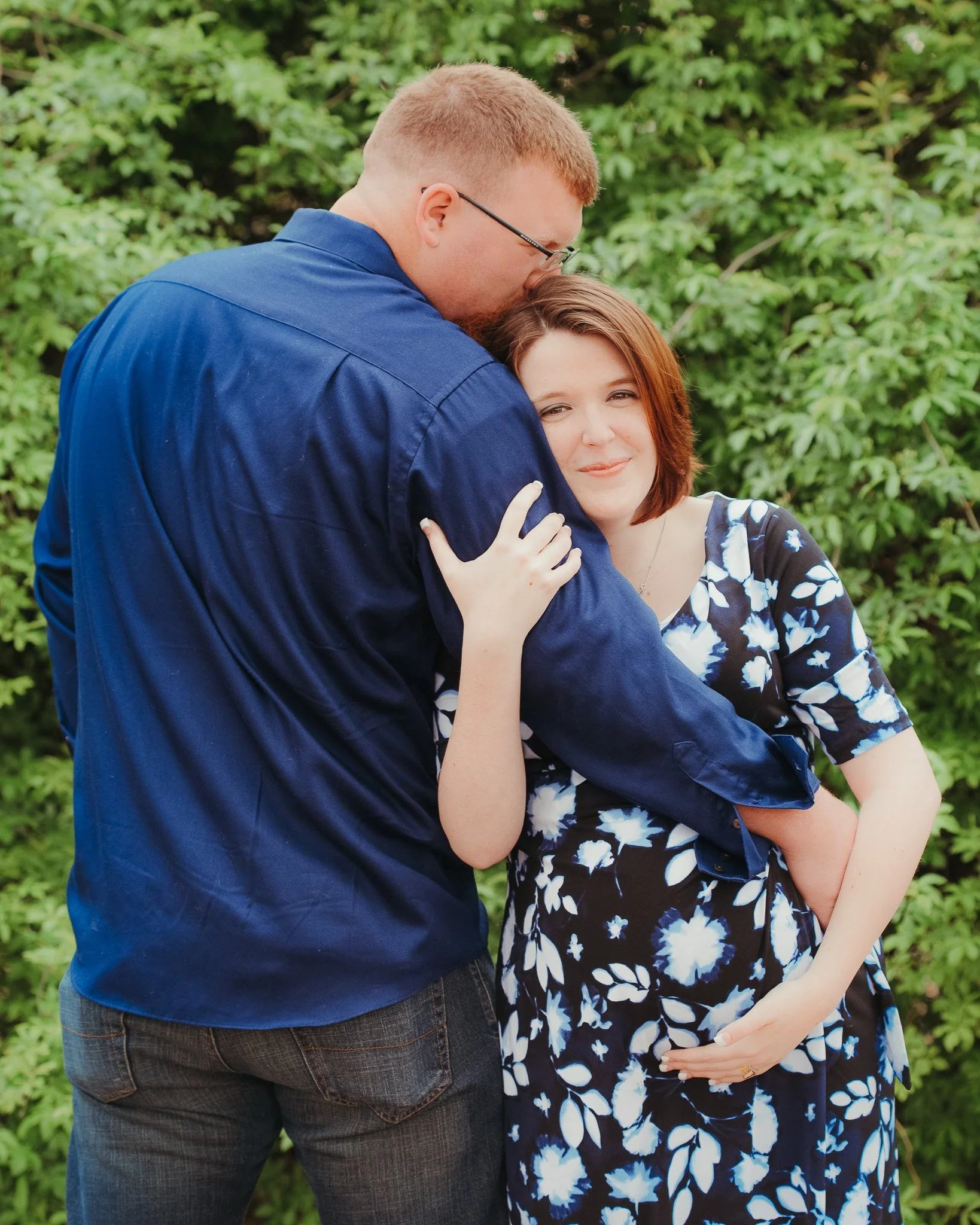 A man and a woman are embracing outdoors, with the man kissing the woman's forehead. The woman has short brown hair and is wearing a black and white floral dress, while the man has short red hair, glasses, and is dressed in a blue shirt and dark jean