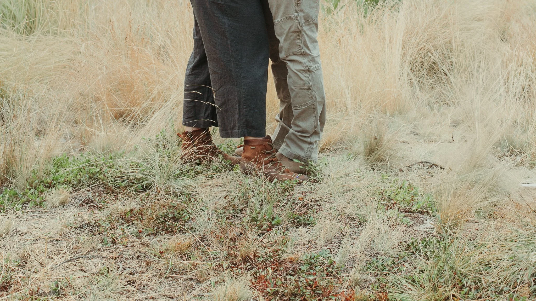Close-up of two people standing outdoors in a grassy, natural setting, showing their legs and feet, wearing hiking boots and casual pants.