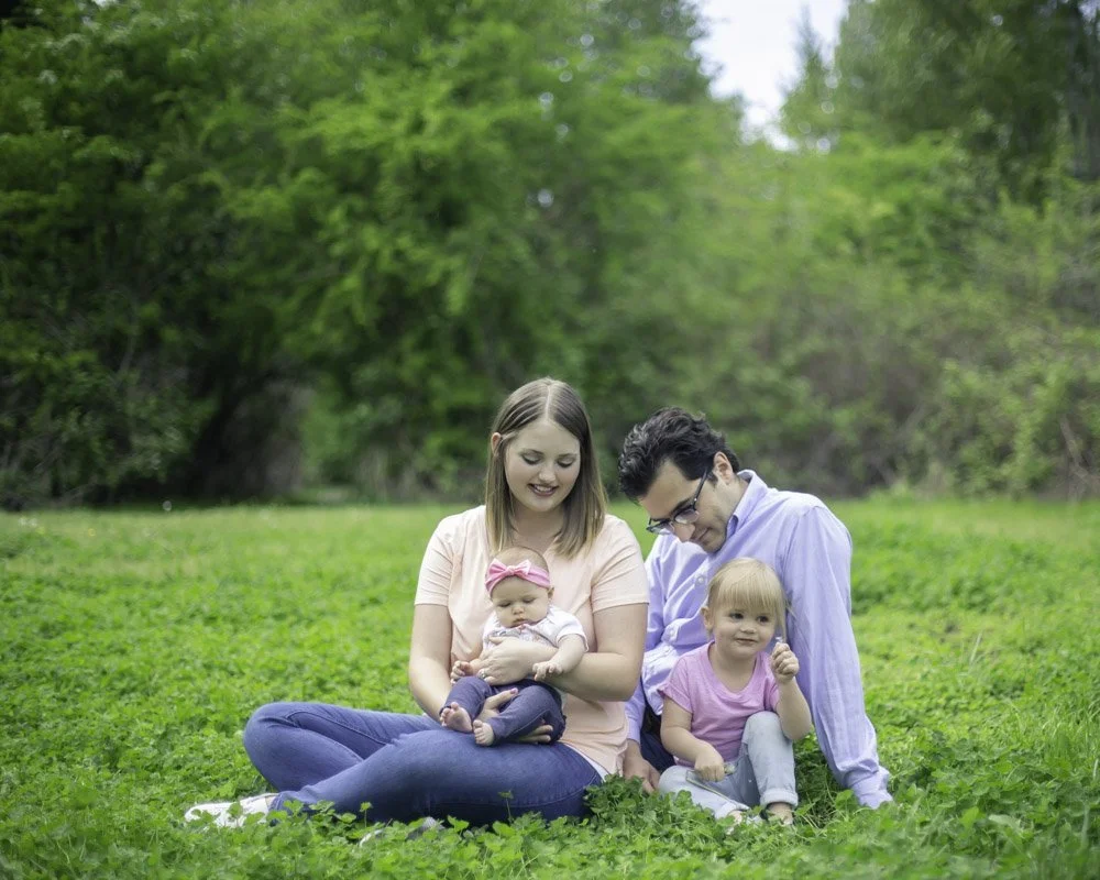 A family of four sitting on grass in a park with trees in the background. The mother holds a baby girl with a pink headband, the father sits next to her with a young girl in a pink shirt, all smiling.