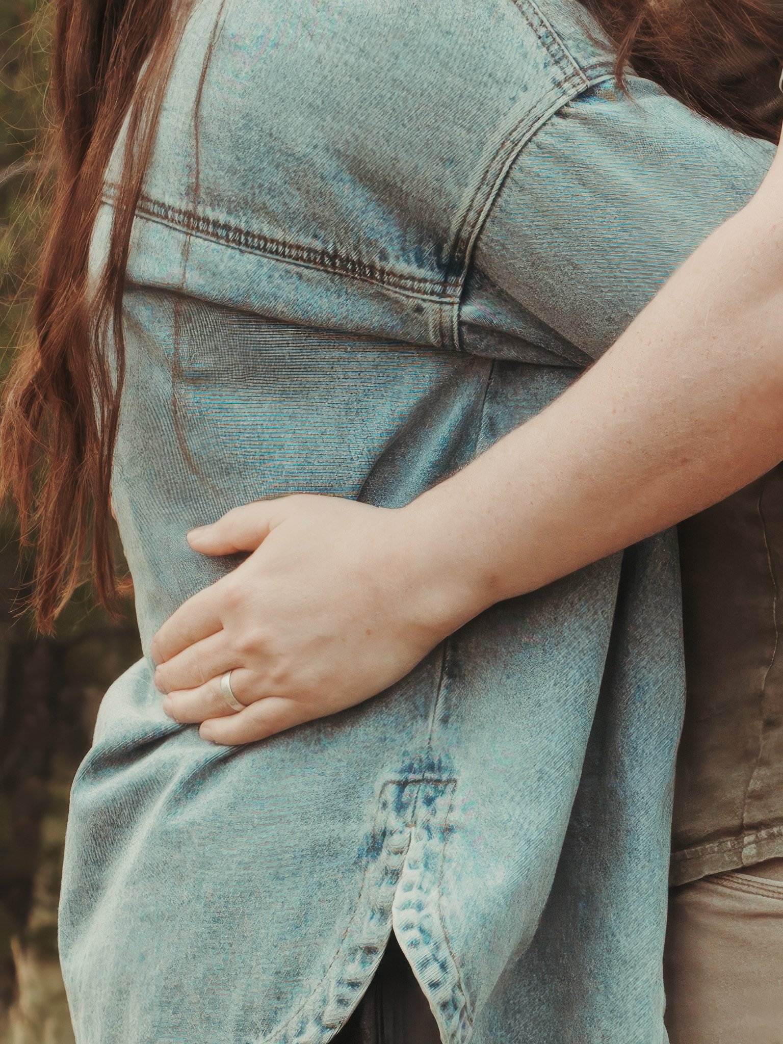 Close-up of a person wearing a light-wash denim jacket and jeans, with long brown hair and a light skin tone. The person is holding their arm across their body, with a blurred outdoor background.