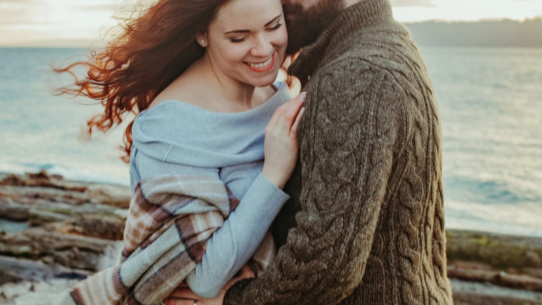 A woman and a man sharing a hug by the water during sunset, with the woman smiling and eyes closed.