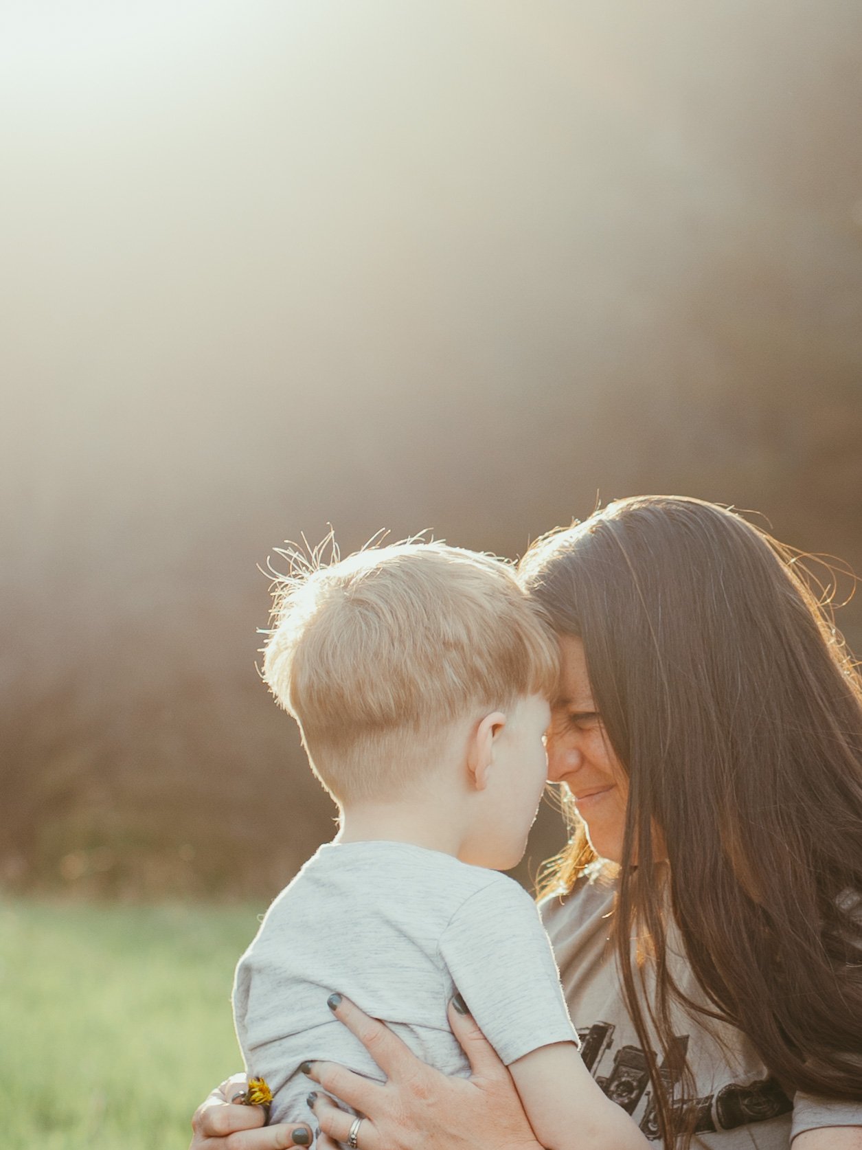 A woman with long dark hair and a young boy with short blond hair are touching foreheads and smiling at each other outdoors during sunset.