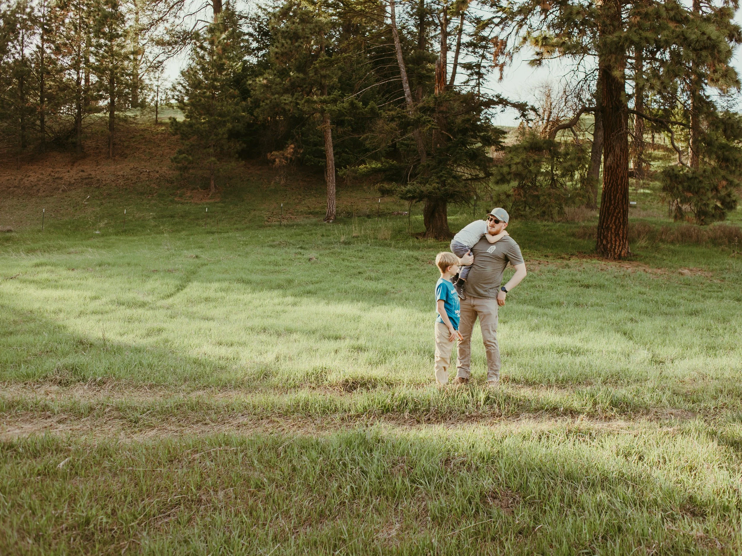 A man with sunglasses and a cap stands on a grassy field with a young child, surrounded by tall trees and a forested background.