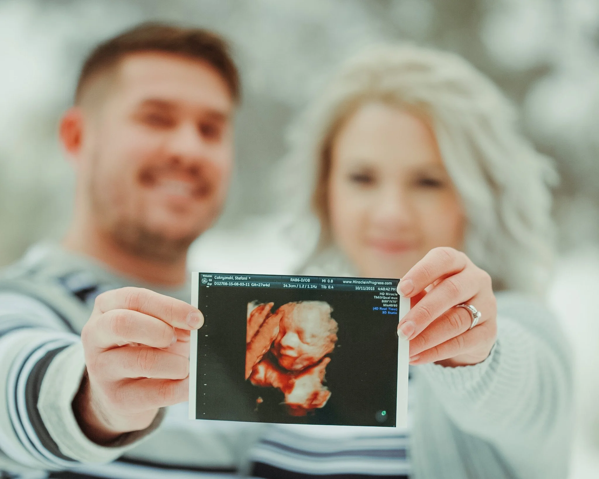 Person holding an ultrasound or medical scan in front of a blurred background with two other people, possibly a couple, smiling.