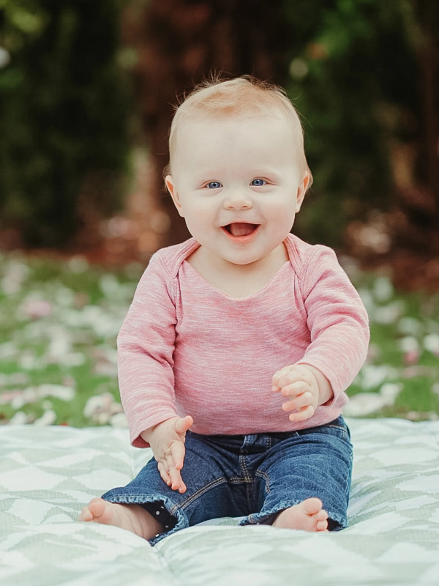 A smiling baby with blue eyes and blonde hair sitting on a blanket outdoors, wearing a pink long-sleeve shirt and denim shorts, with blurred greenery in the background.