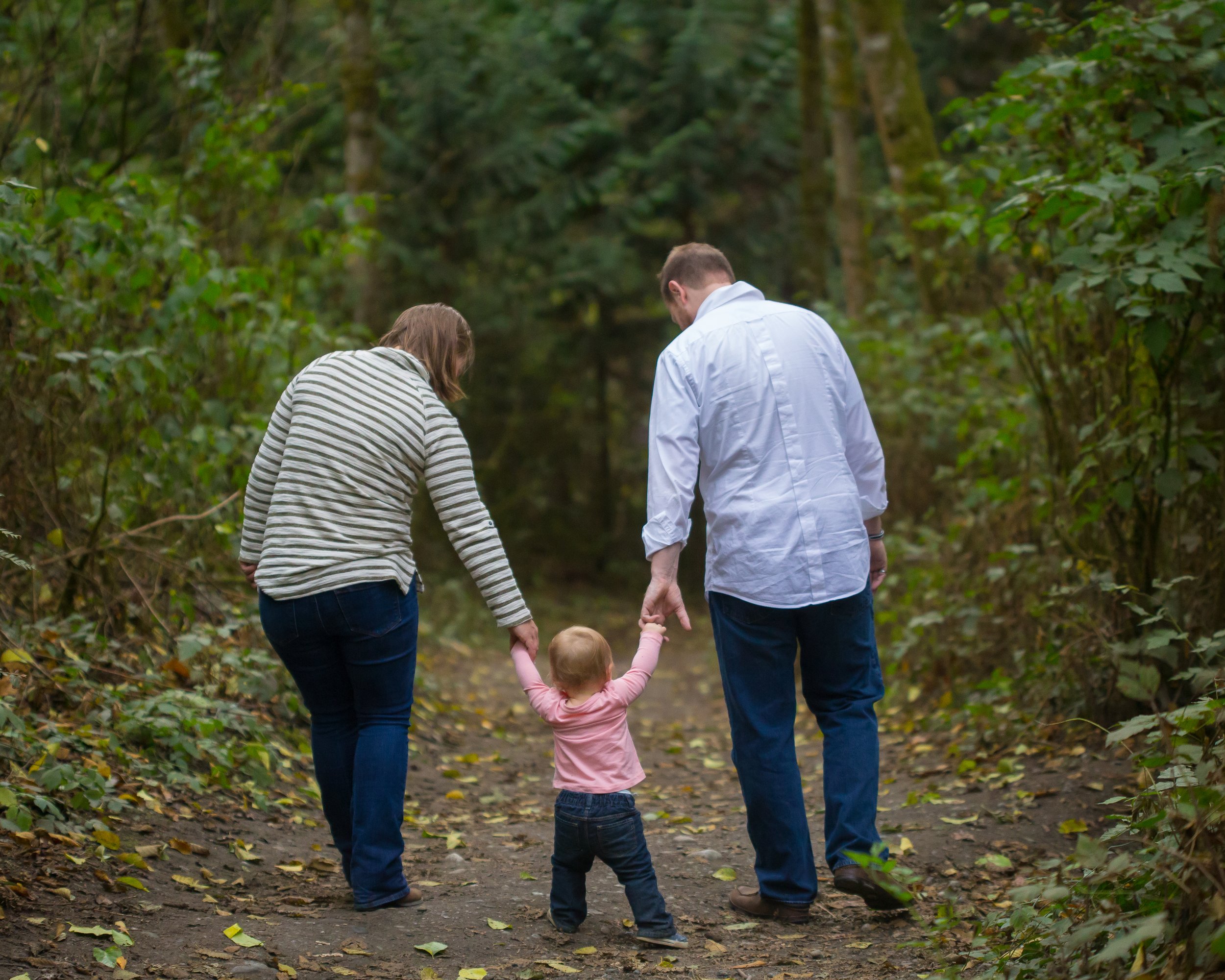 A family of three walking on a wooded trail, holding hands. The adults are dressed in casual clothing, and the child is a young girl in a pink jacket.