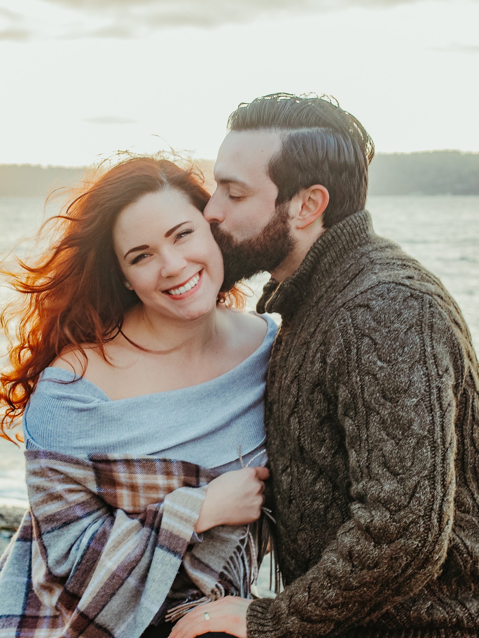 A happy woman with red hair smiling as a man kisses her on the forehead near a body of water, likely during sunset or sunrise.