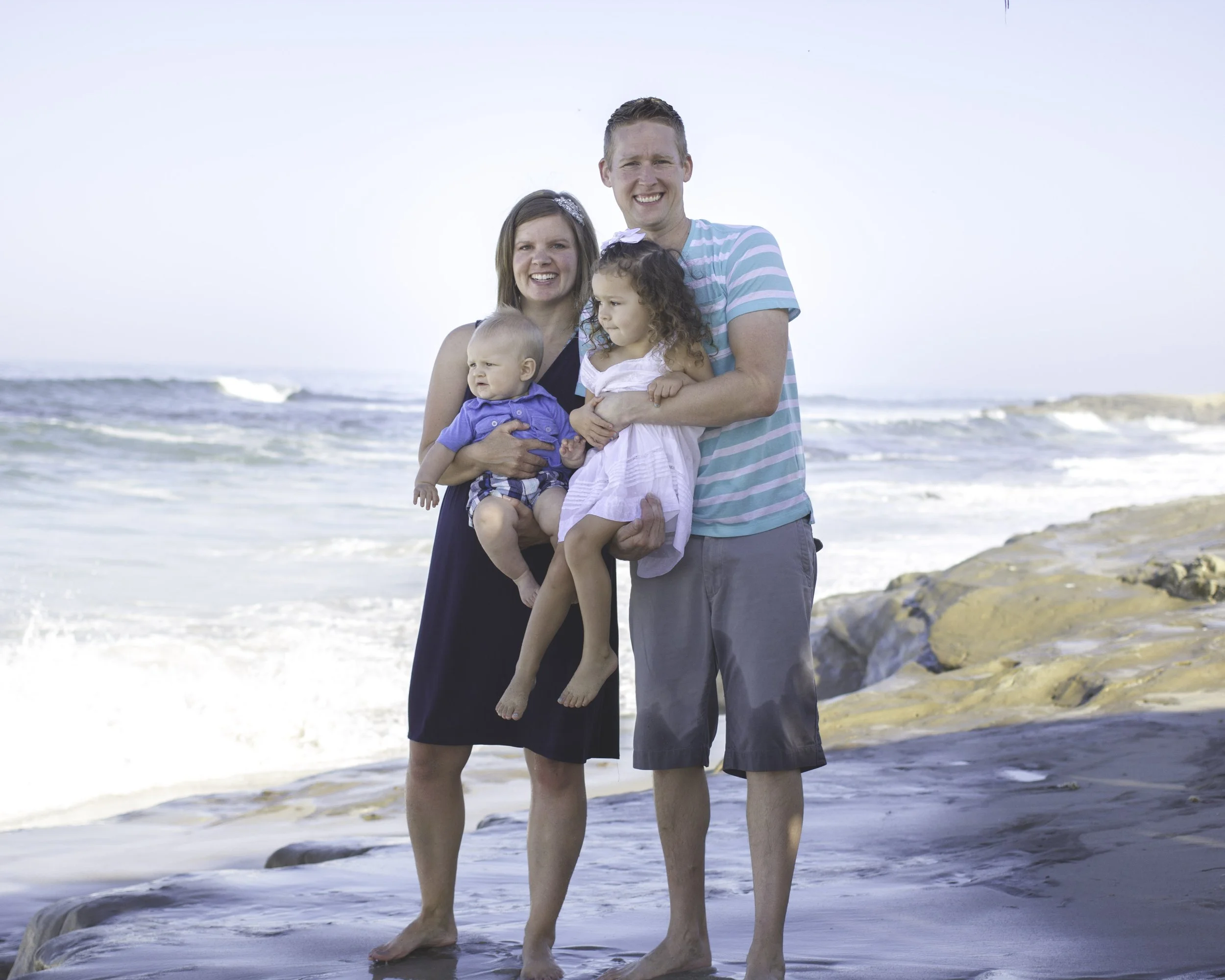 A family of four standing on a beach, smiling at the camera, with ocean waves and a rocky shoreline visible in the background.