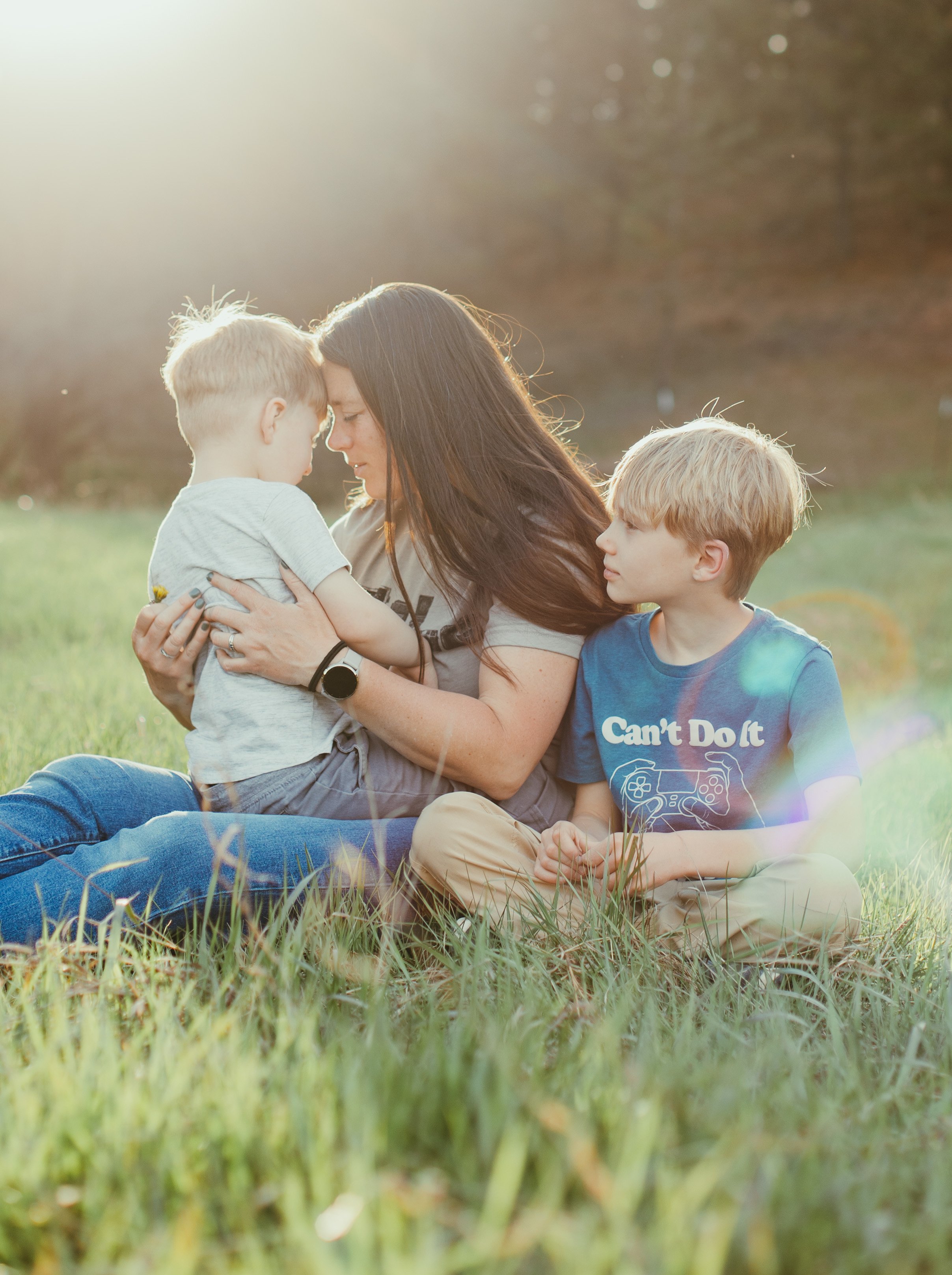 A woman with long dark hair holding a young boy with blond hair and a second boy with blond hair wearing a blue shirt sits on grass in a field at sunset. The woman and the boy she is holding share a forehead-to-forehead moment. The second boy looks o