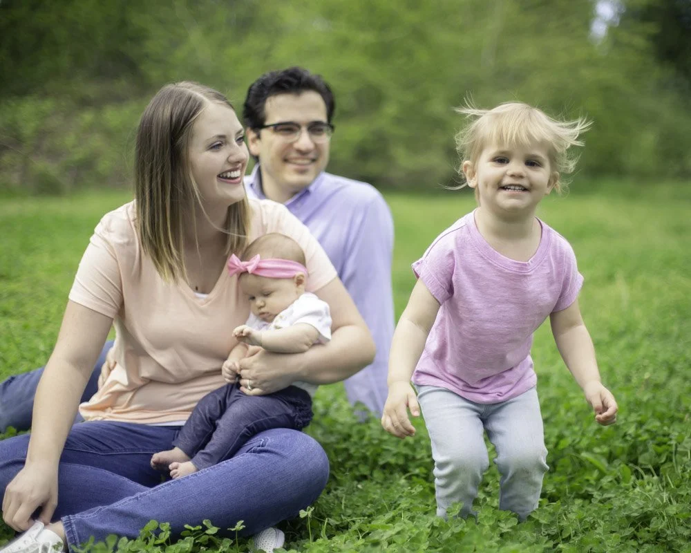 Family of four sitting and playing on grassy field with trees in the background, smiling and enjoying outdoor time.