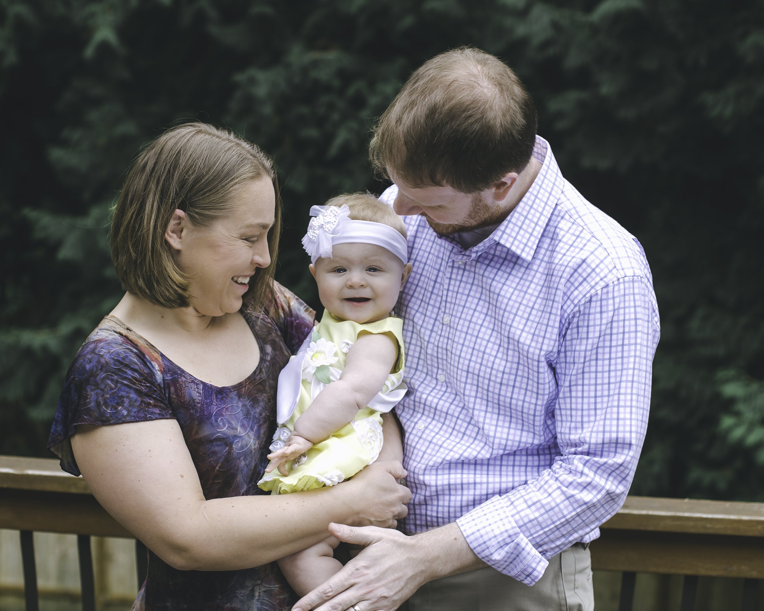 A family of three, including a woman, a man, and a baby girl, outdoors with trees in the background. The woman is smiling and holding the baby, while the man is bending down close to them, all sharing a happy moment.