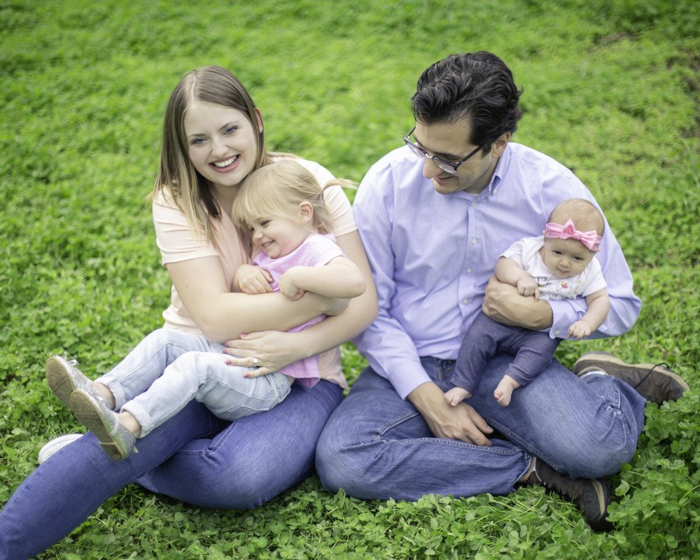 A family of four sitting on the grass outdoors, smiling and playing together. The mother is holding a young girl, and the father is holding a baby girl with a pink bow in her hair.