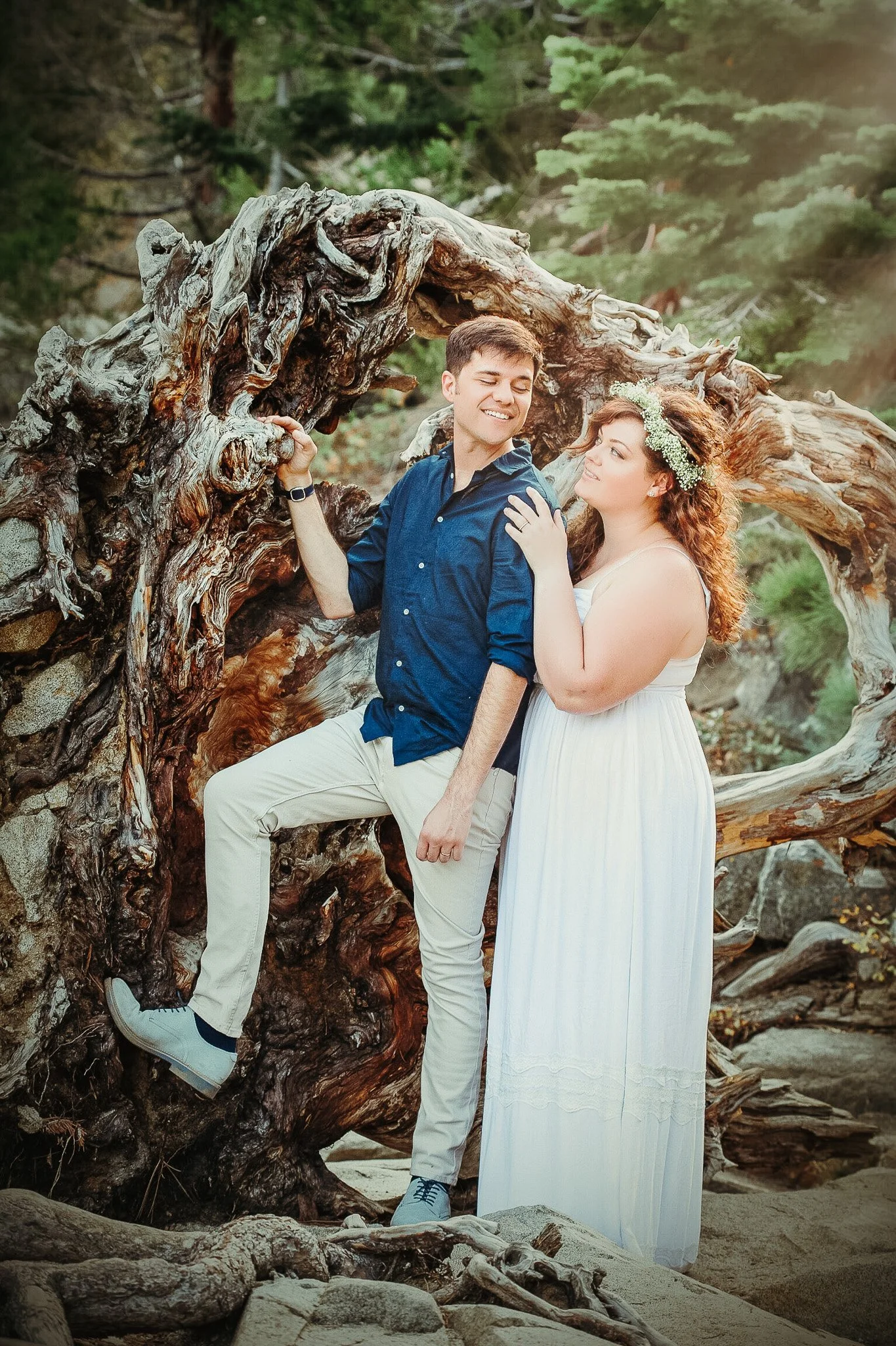 A young man and woman stand close to each other near a large, weathered tree trunk outdoors. The man wears a blue shirt and beige pants, smiling with eyes closed, while the woman, with curly hair and a flower crown, looks at him, touching his shoulder. The setting appears to be a natural, forested area with rocks and sandy ground.