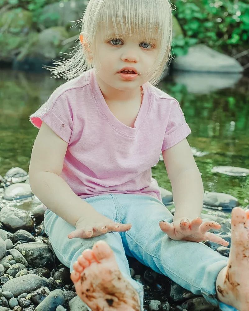 A young girl with blonde hair and blue eyes sitting by a river on a rocky shoreline, wearing a pink shirt and light blue pants, with dirt on her hands and feet.