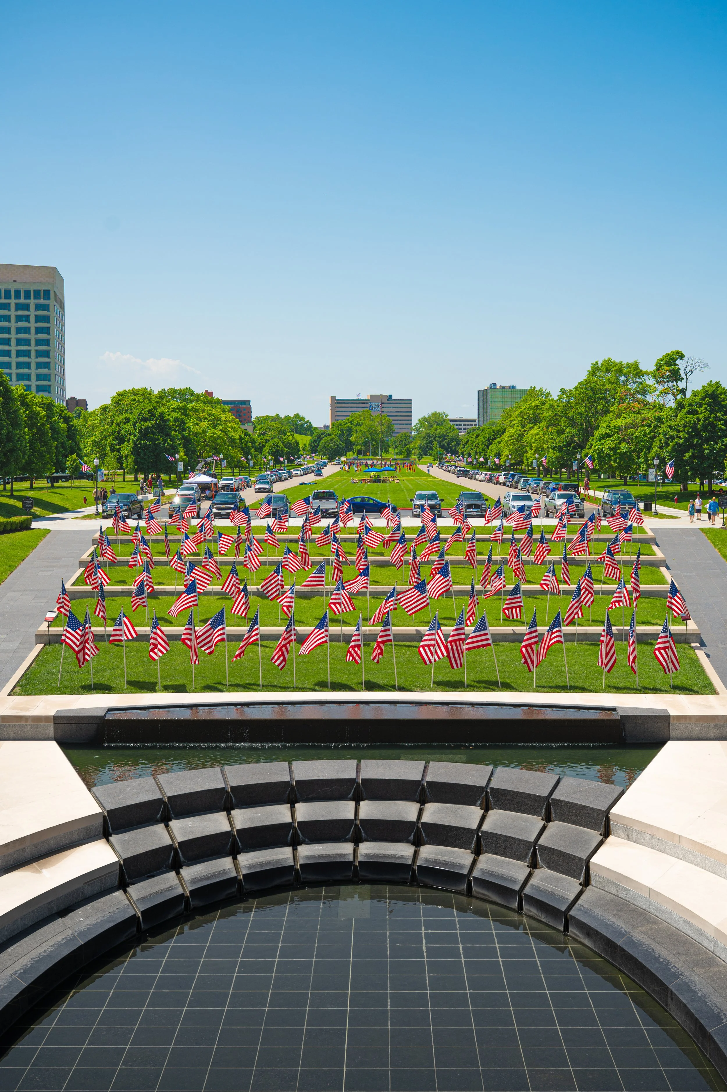 Memorial with American flags arranged in a grid, reflecting in a water feature, on a bright day with clear blue sky, tall buildings and trees in the background.
