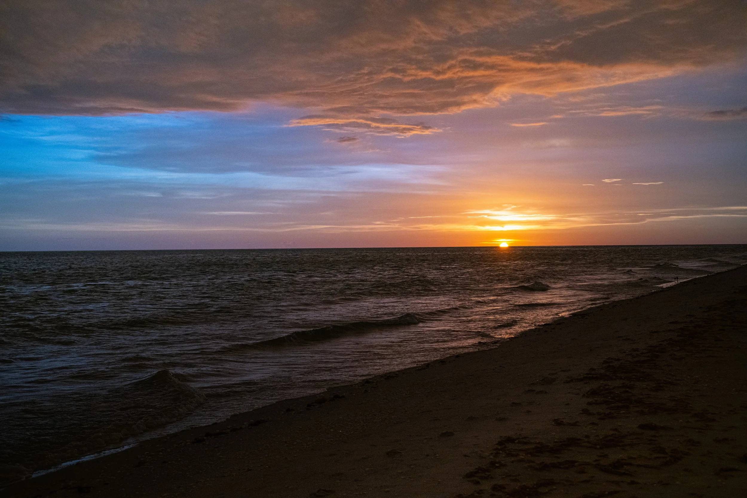 A sunset over the ocean with a partly cloudy sky, the sun near the horizon casting a golden glow, and waves gently crashing on a beach.
