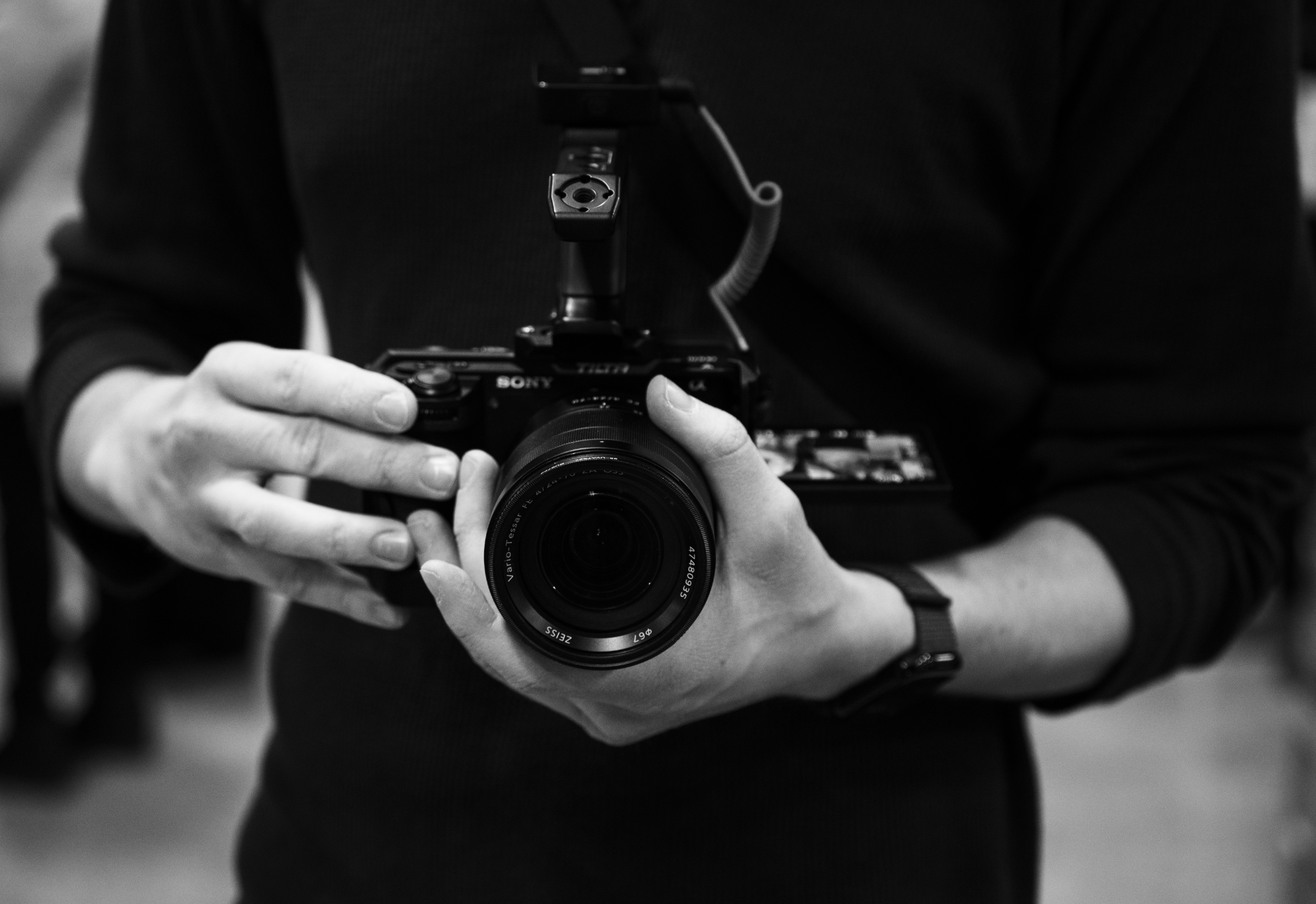 Black and white photo of a person holding a camera, viewfinder facing the mirror, person wearing a dark long-sleeve shirt, and a strap attached to the camera visible around the neck.