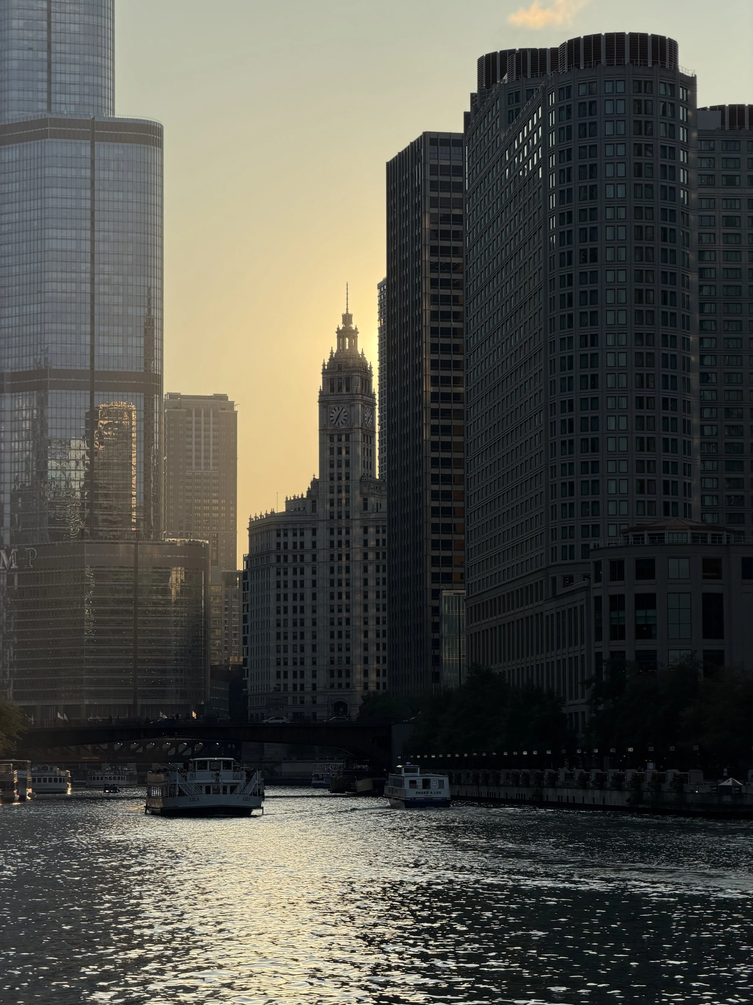 City skyline with tall modern buildings along a river at sunset, with boats on the water and a clock tower in the background.