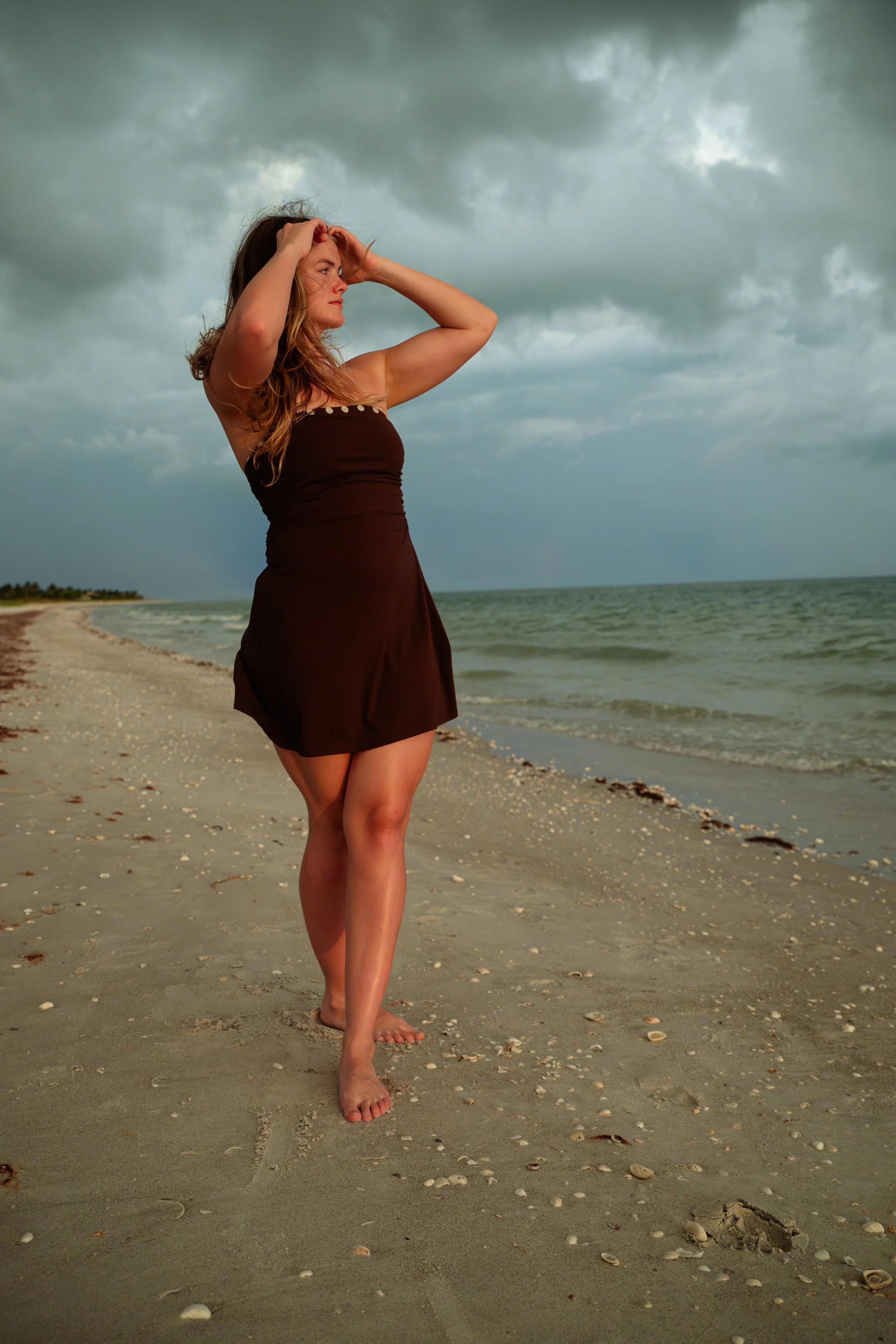 A woman standing barefoot on a sandy beach with seashells scattered around, wearing a dark strapless dress, looking to the left with her hands on her head under a cloudy sky.