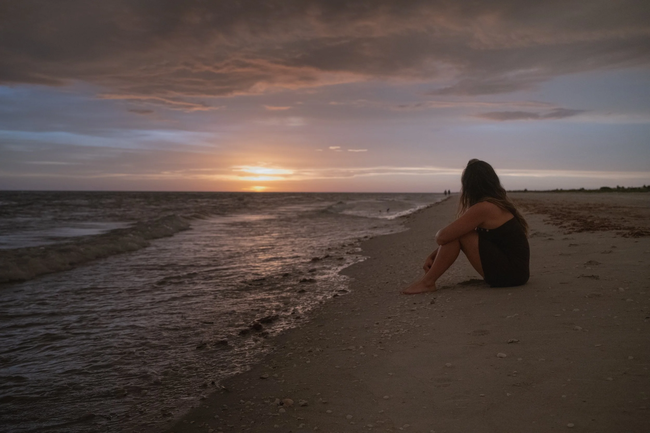 A woman sitting alone on the beach near the shoreline at sunset, looking out toward the distant horizon with clouds in the sky.