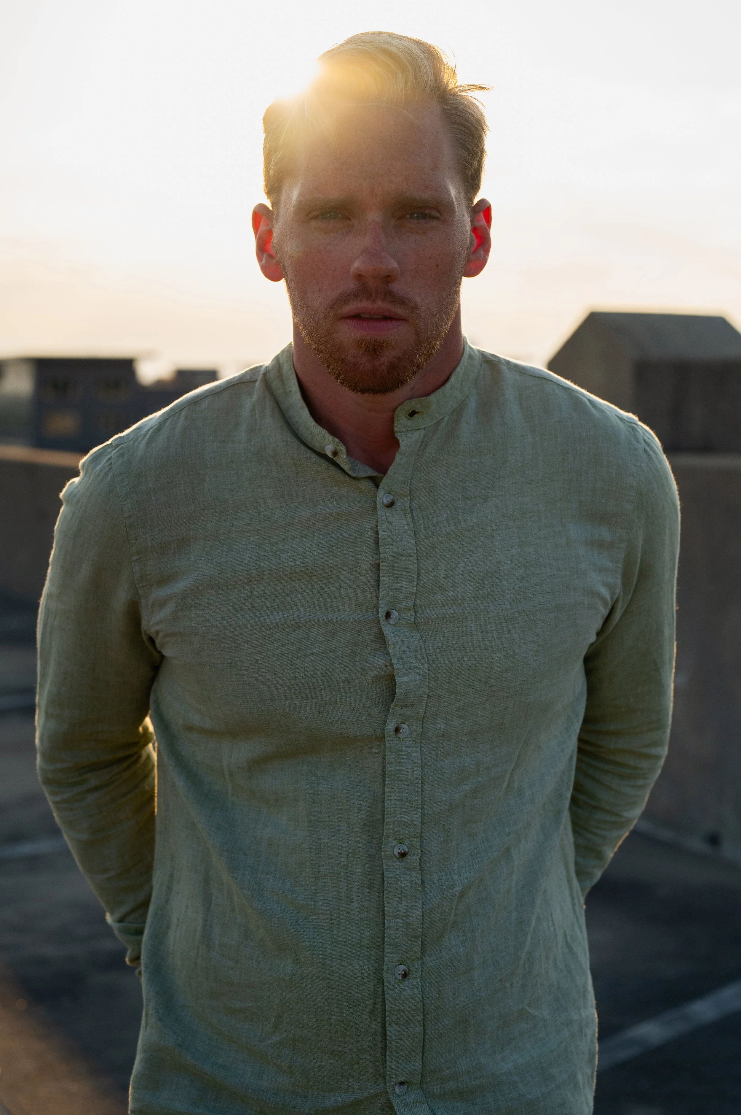 A man with short blonde hair, beard, and light-colored shirt standing outdoors on a rooftop with the sun setting behind him. The sky is bright, casting a warm glow on his face.