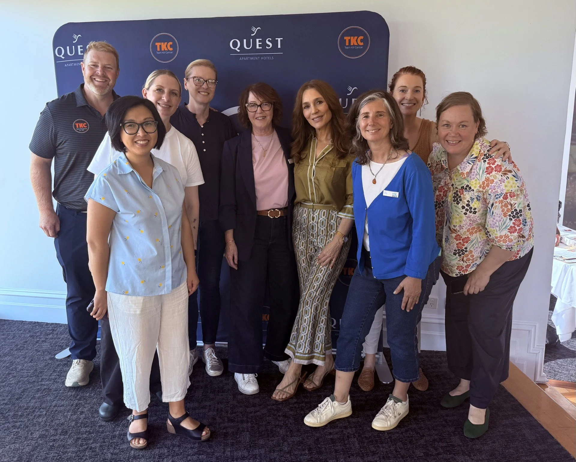 A group of nine diverse people, smiling, standing in front of a blue QUEST banner with logos, in a well-lit room, posing for a photo at an event.