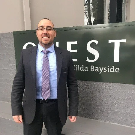 A man in a suit standing outside a building with a sign that says 'GUEST' and 'Brida Bayside'.