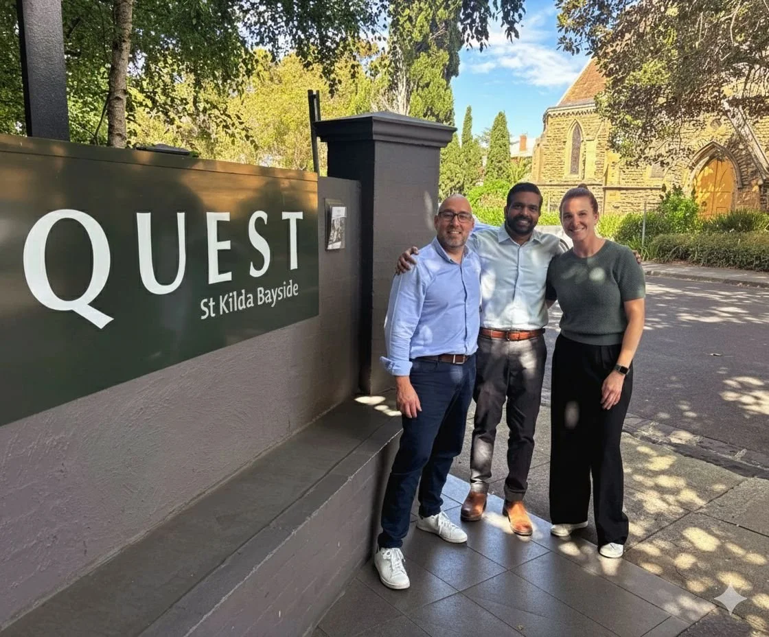Three people standing closely together outdoors near a sign that reads 'Quest St Kilda Bayside.' There is a church or historic building in the background, surrounded by trees and sunlight.