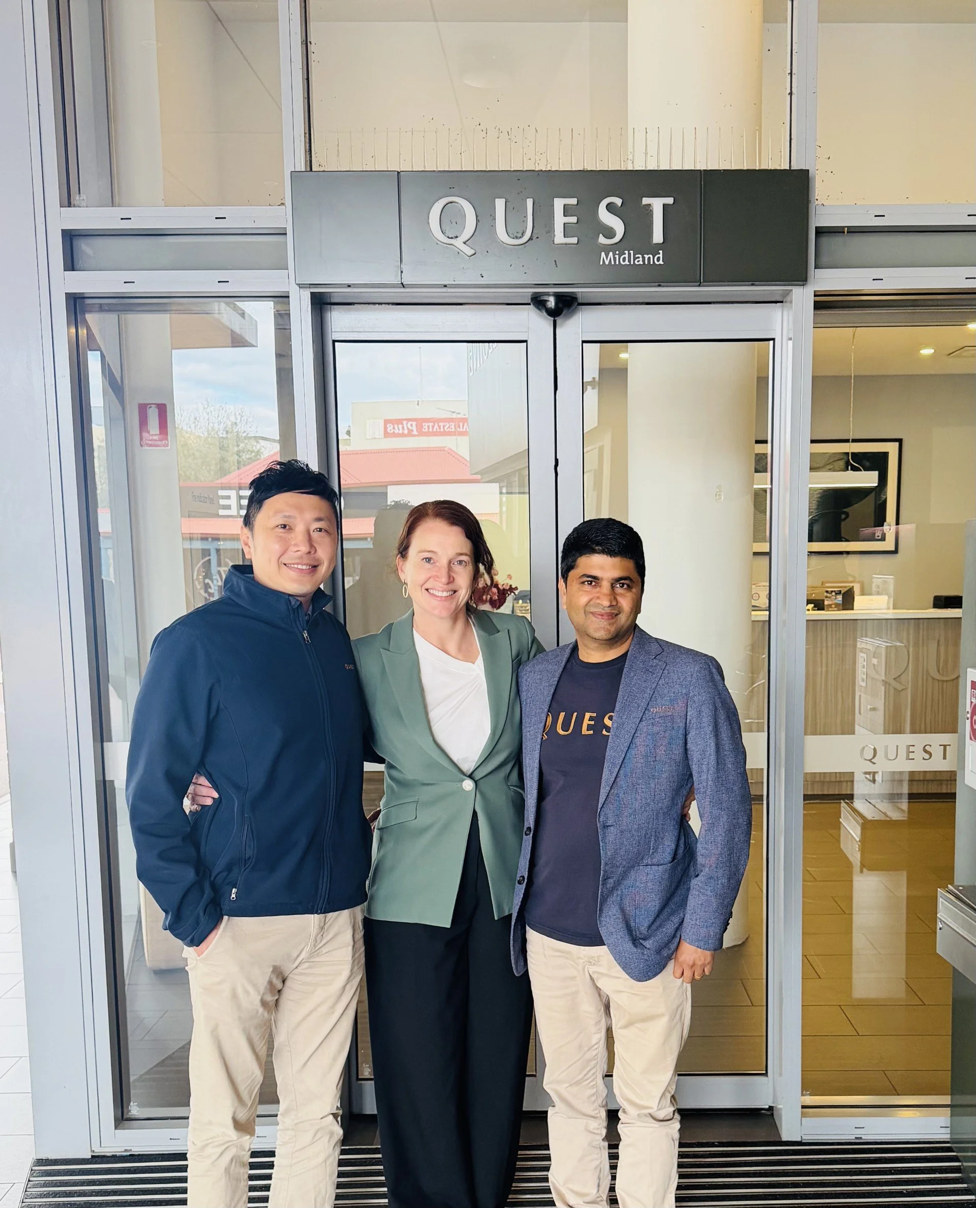 Three people standing in front of a Quest Midland sign, smiling, inside a building with glass doors and windows, with a reflection of outdoor buildings and trees.