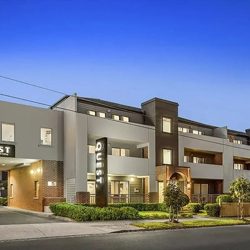 Modern apartment building with a sign reading 'QOVEST' near the entrance, illuminated at dusk, with bushes and small trees in front.