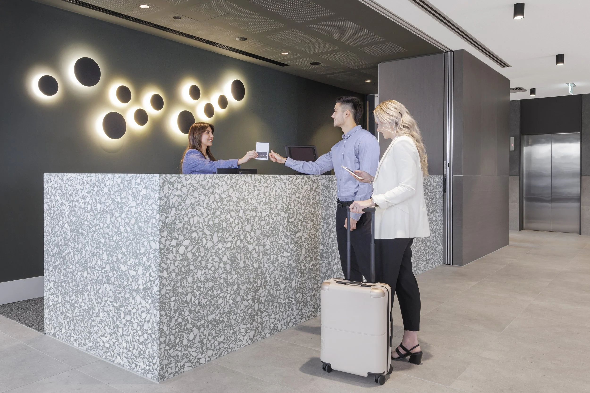 A woman checks into a hotel at the reception desk with two guests, one man and one woman, waiting in line with luggage. The woman checking in is handing over a card to the receptionist, and the woman with luggage is looking down at her phone.