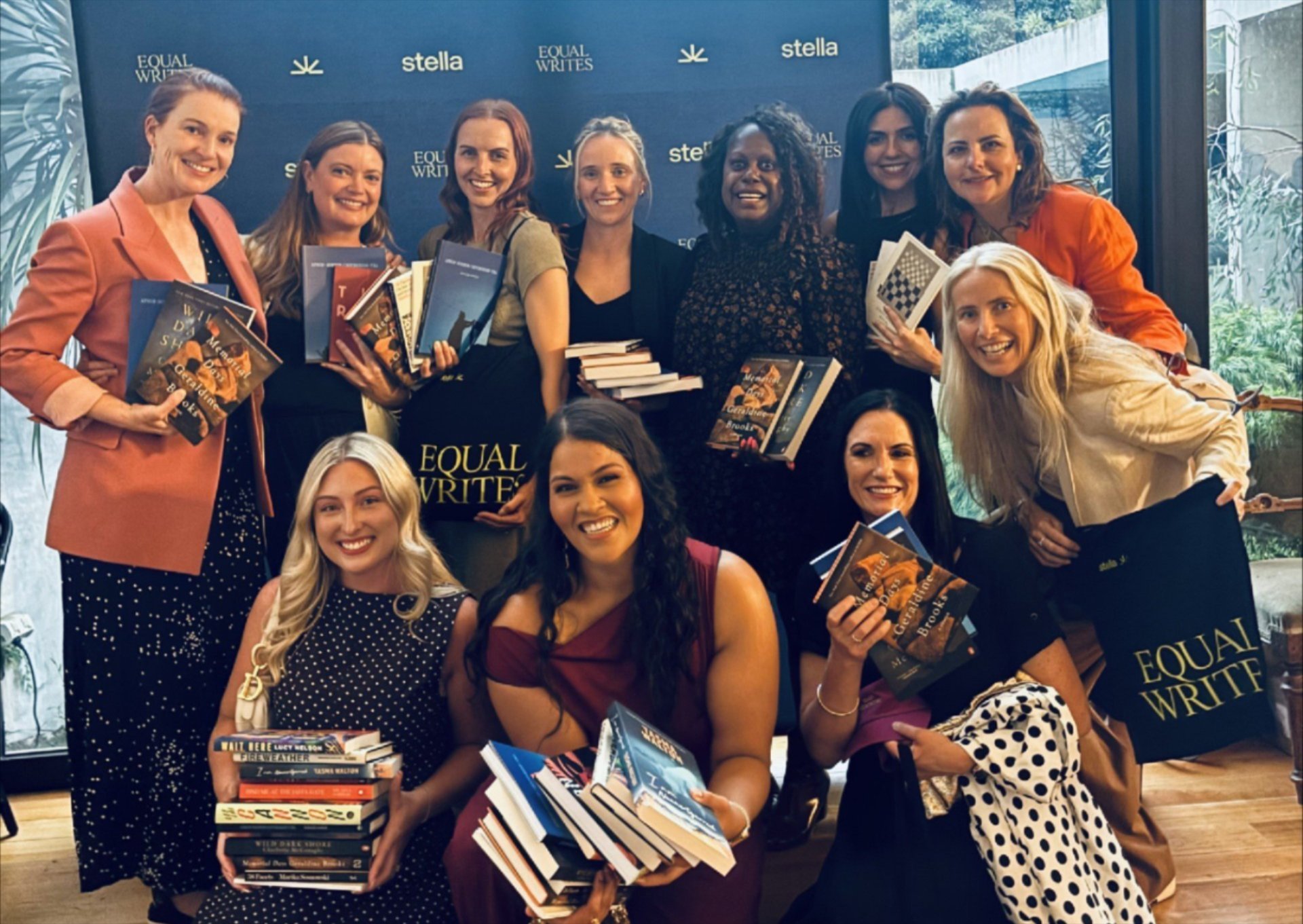 Group of women at a book event, holding stacks of books, standing in front of a backdrop that says 'Equal Writes' and 'Stella'.