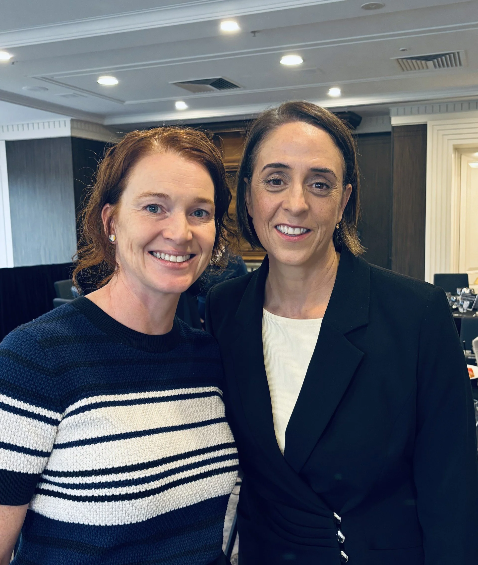 Close-up photo of two women smiling, standing close together indoors. One woman has red hair and is wearing a navy blue and white striped sweater. The other woman has brown hair and is wearing a black blazer over a white top.