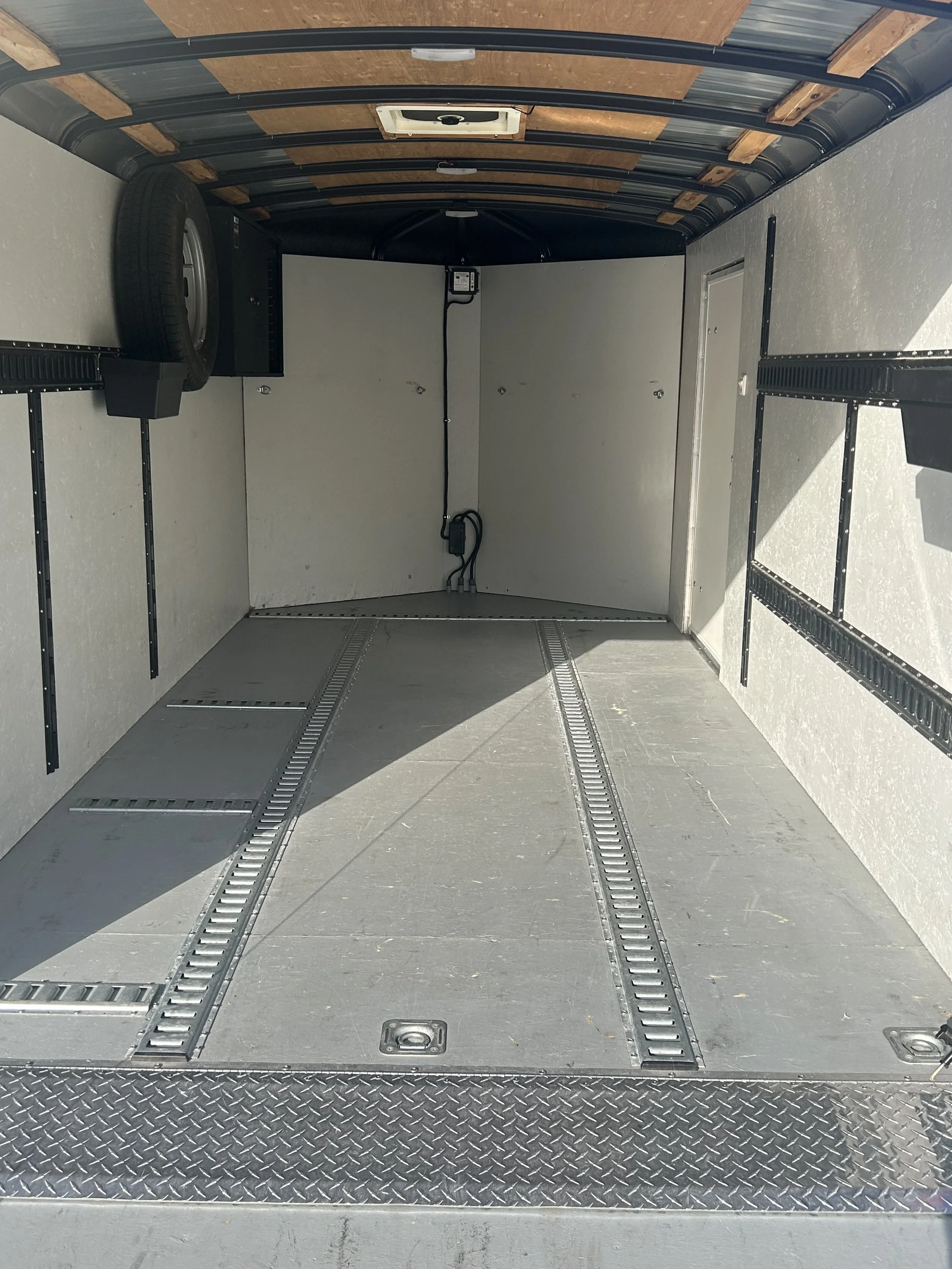 Inside of an empty silver enclosed trailer with a tire mounted on the left side and black beams with wooden slats on the ceiling, along with wiring and electrical components.