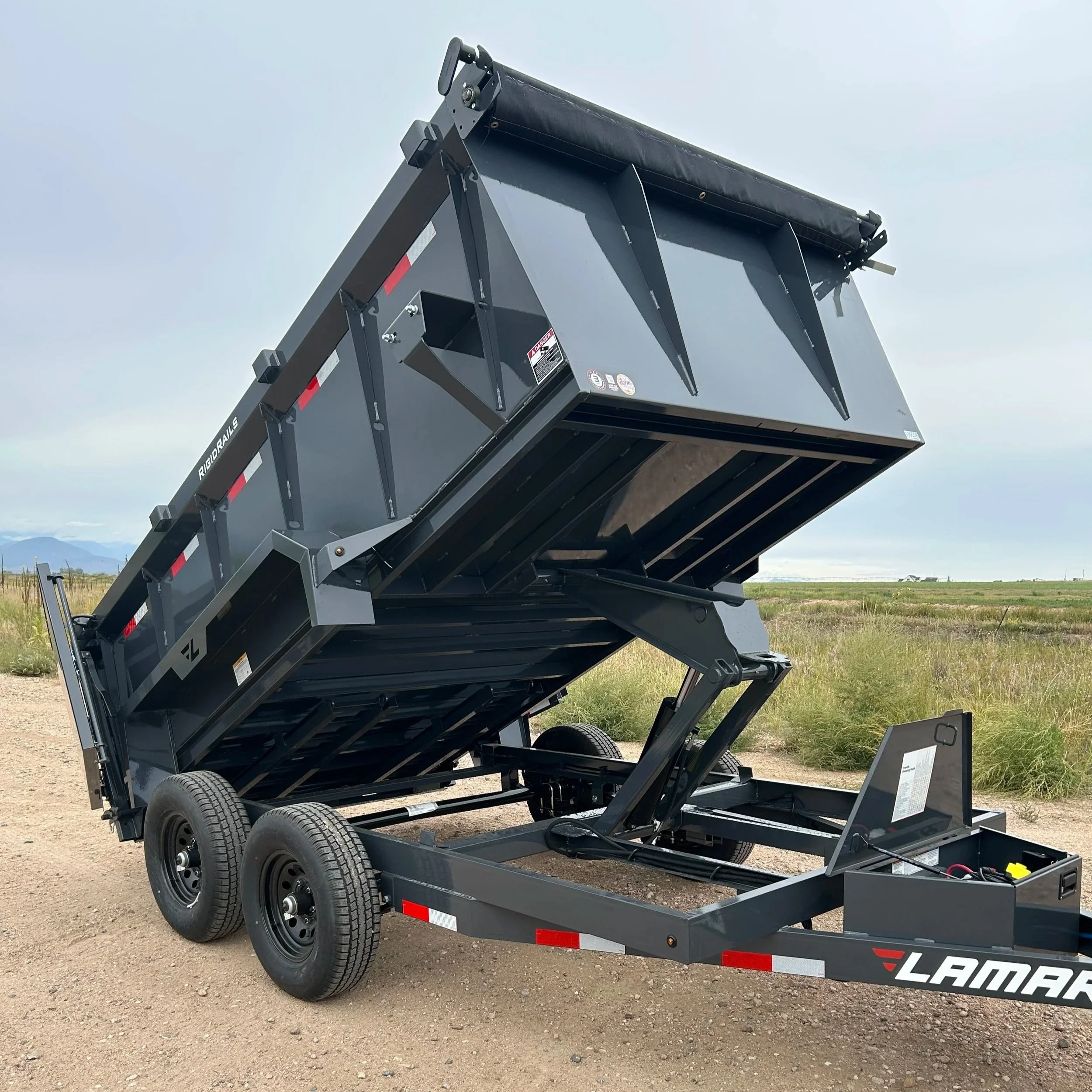 A dump trailer, showing a hydraulic lift mechanism, parked on a dirt field with mountains in the background.