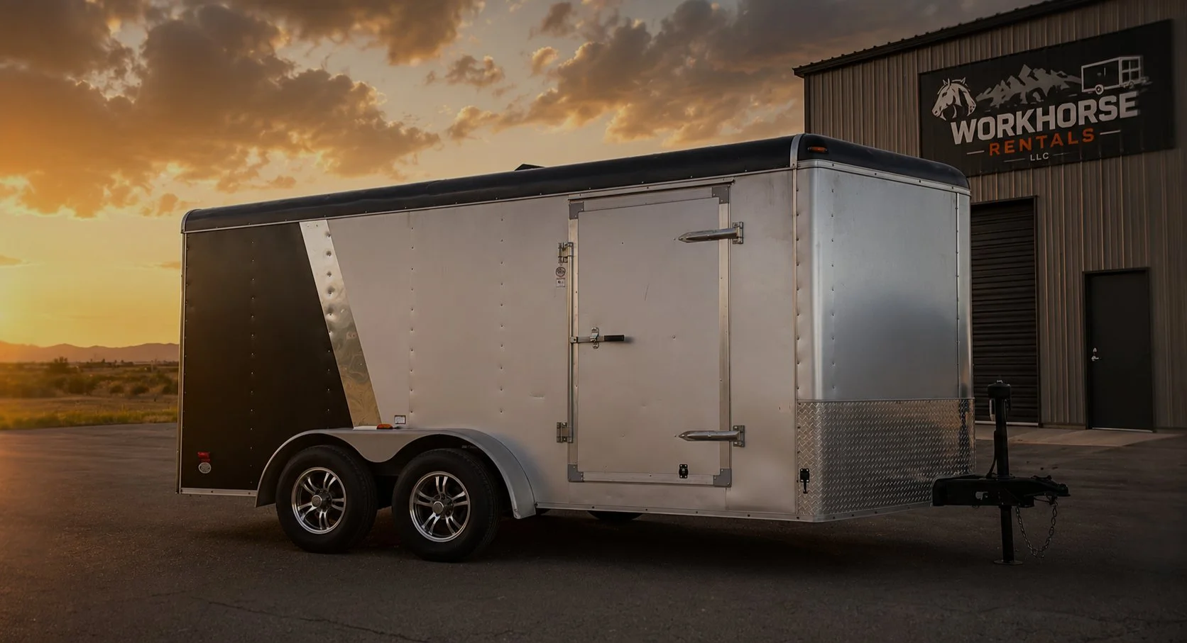 A silver and black enclosed trailer parked outside a building with a "Workhorse Rentals LLC" sign, during sunset with mountains in the background.